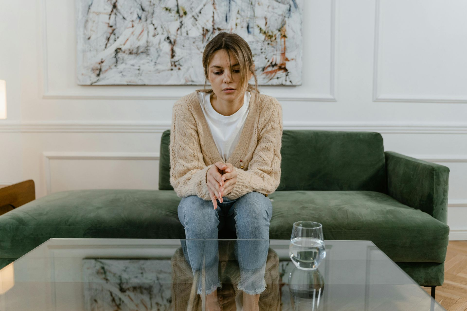 Woman sits on a green couch, looking down with a sad expression. A glass of water sits on a table.