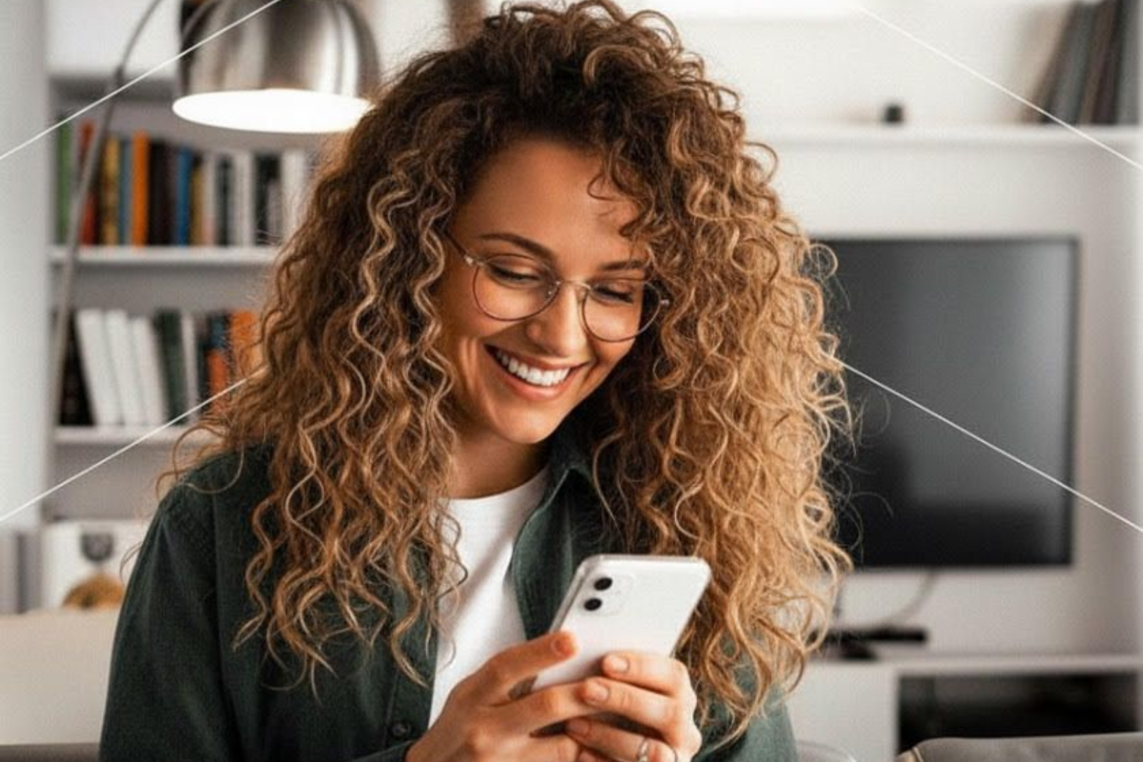 Une femme aux cheveux bouclés sourit en regardant son téléphone dans un salon.