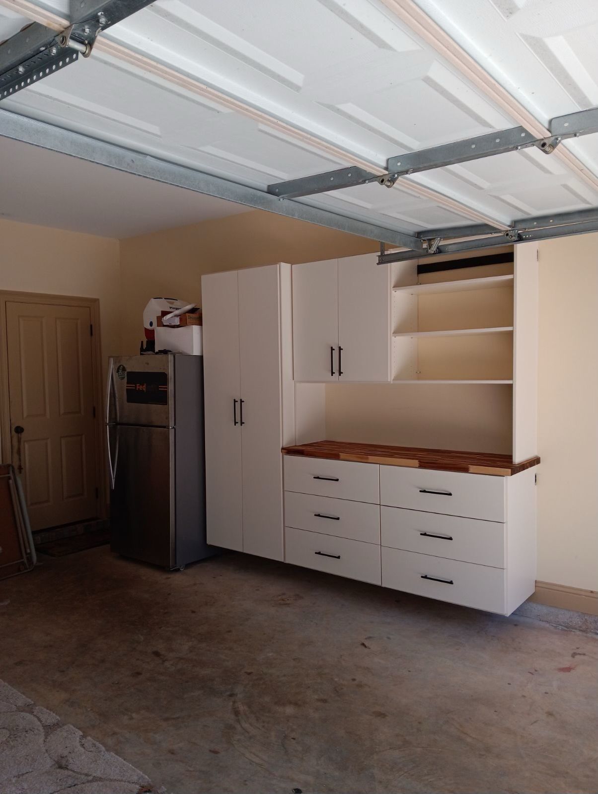White cabinets with a countertop and drawers mounted on a garage wall, positioned beside a silver refrigerator.