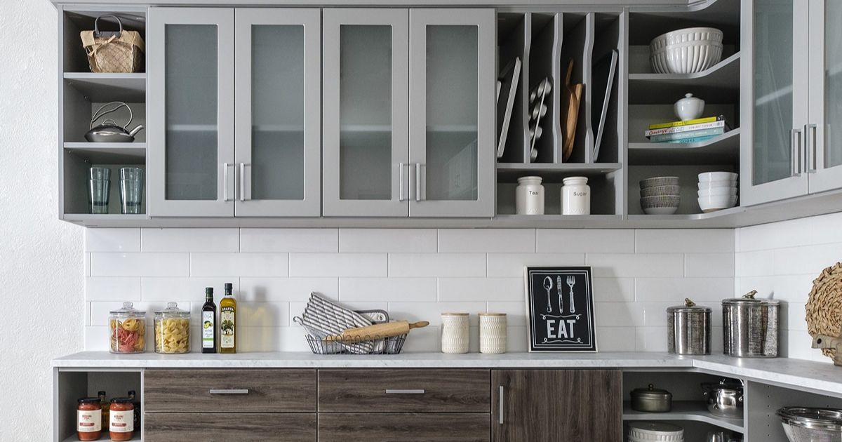 Modern kitchen with gray and brown cabinets, white tile backsplash, and countertop items. Upper cabinets have glass doors and open shelves.
