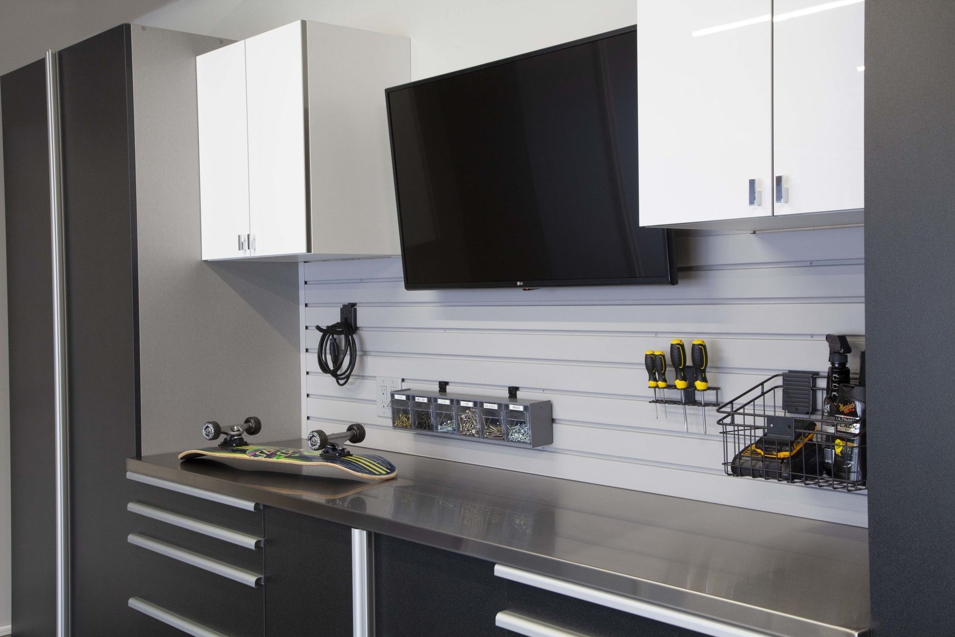 Garage workspace with cabinets, TV, and tools on a slat wall. Black and white color scheme, stainless steel countertop.