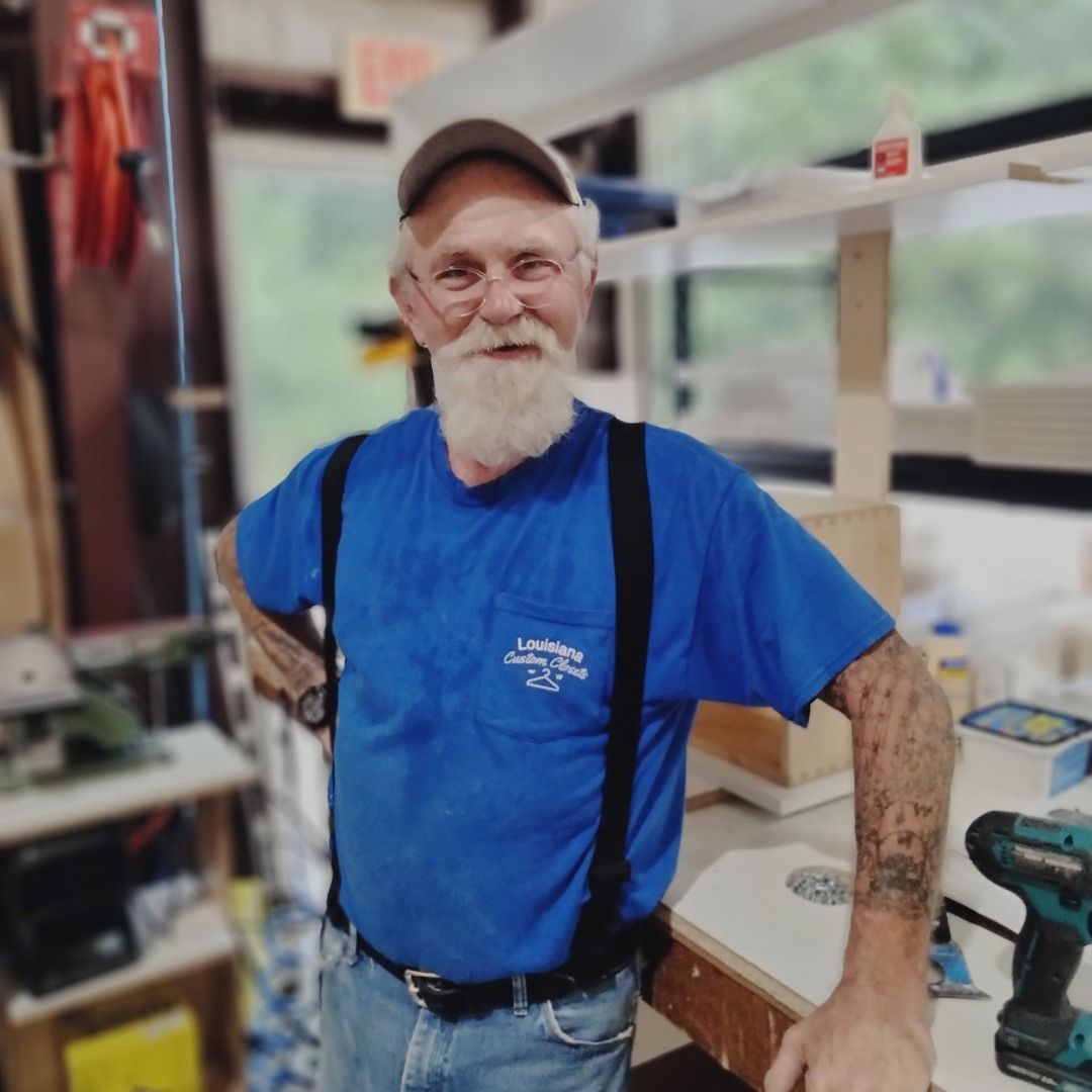 Man with white beard wearing a blue shirt and suspenders in a workshop, smiling.