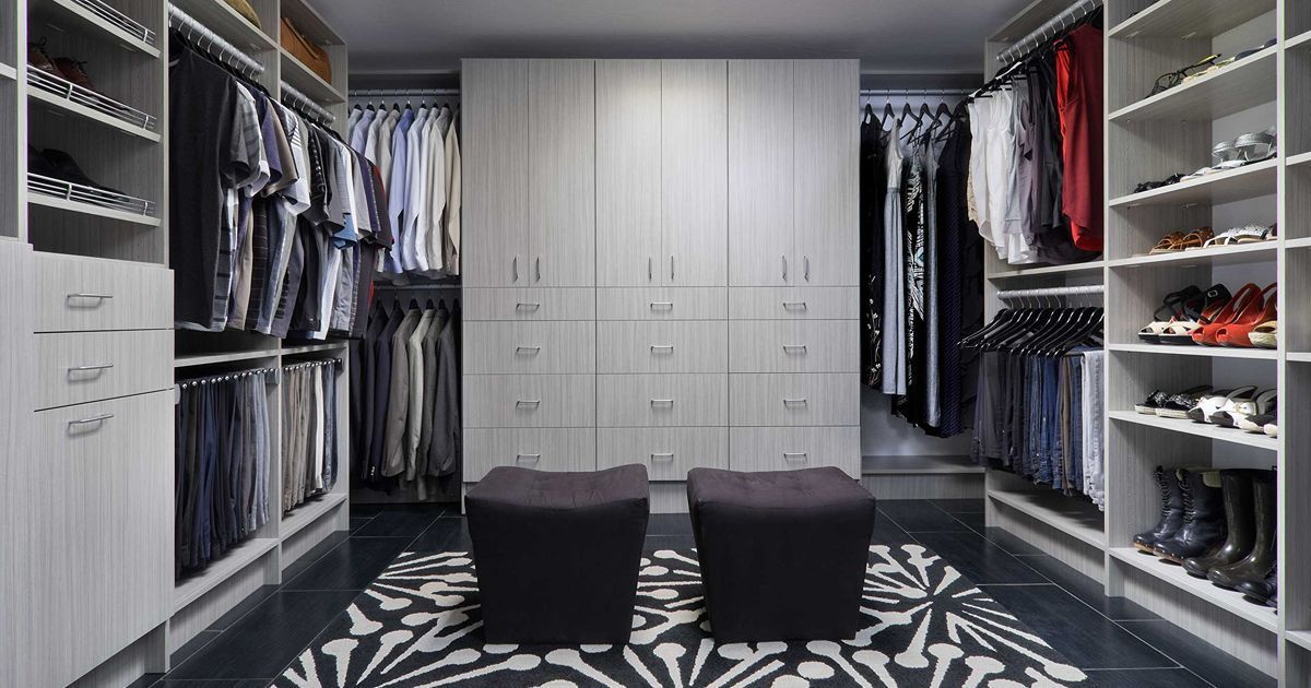 Walk-in closet with gray cabinetry, hanging clothes, and shoe shelves. Two black ottomans sit on a patterned rug.