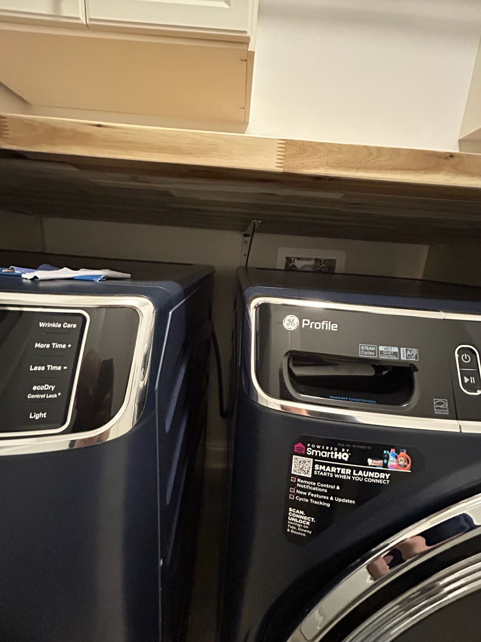 Two navy blue washing machines side-by-side in a laundry room with a shelf above them.