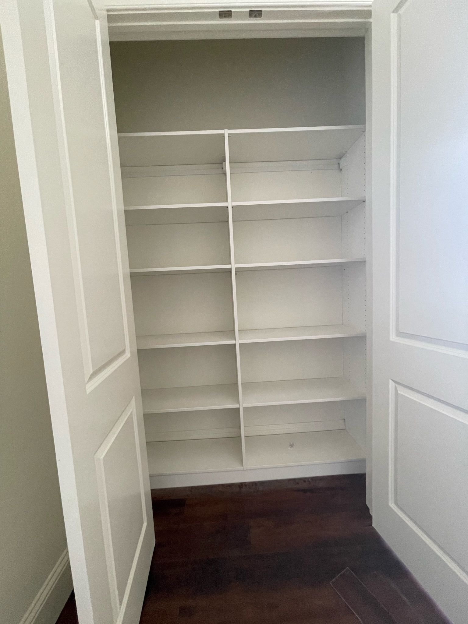 Empty white pantry with multiple shelves, open white doors, and dark wood floor.