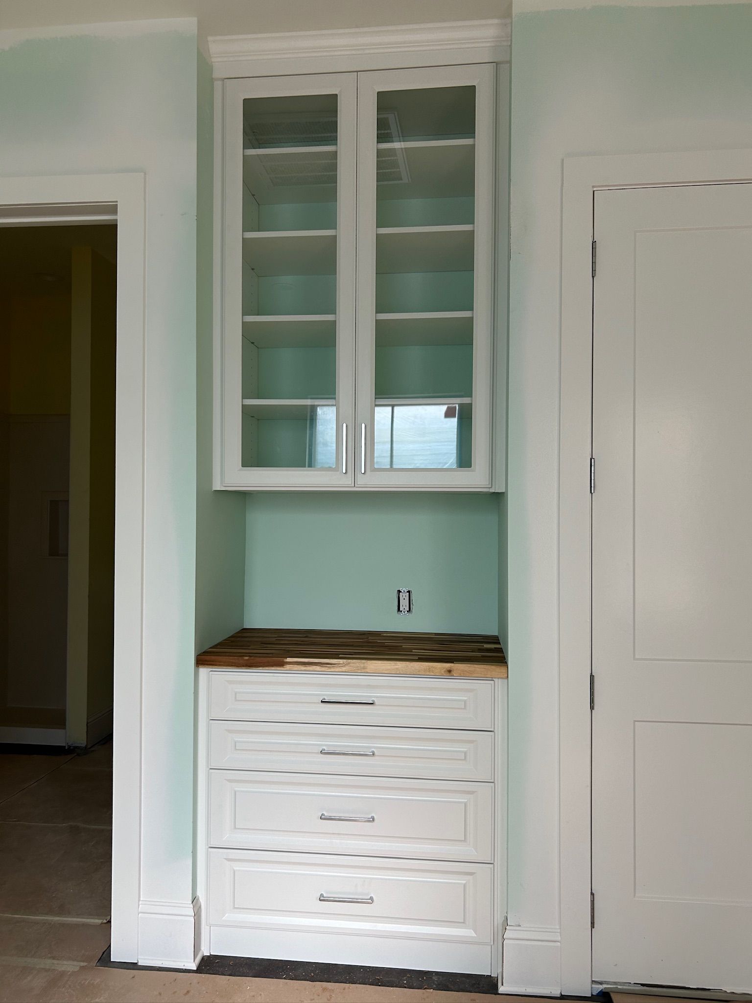 Built-in white cabinet unit with glass-door upper cabinets, drawers, and wood counter against a mint wall.