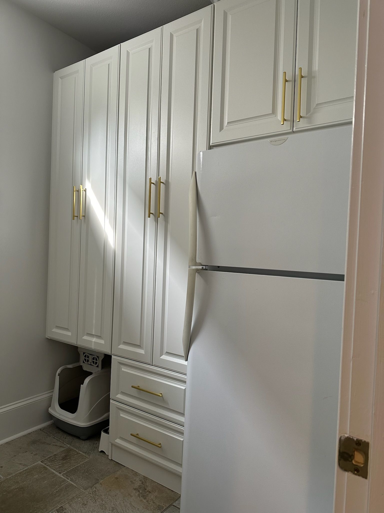 White cabinets with gold handles, refrigerator, and a cat litter box in a small room.