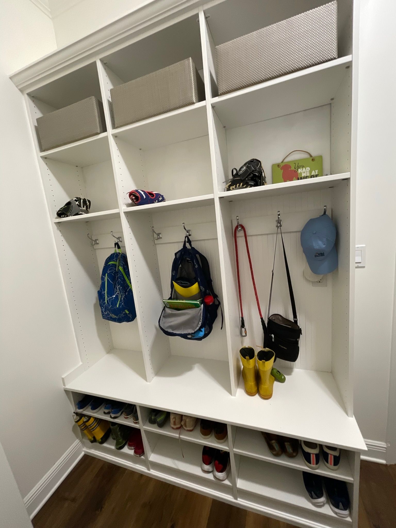 White built-in storage unit with shelves and hooks. Backpacks, shoes, and bins are stored inside.