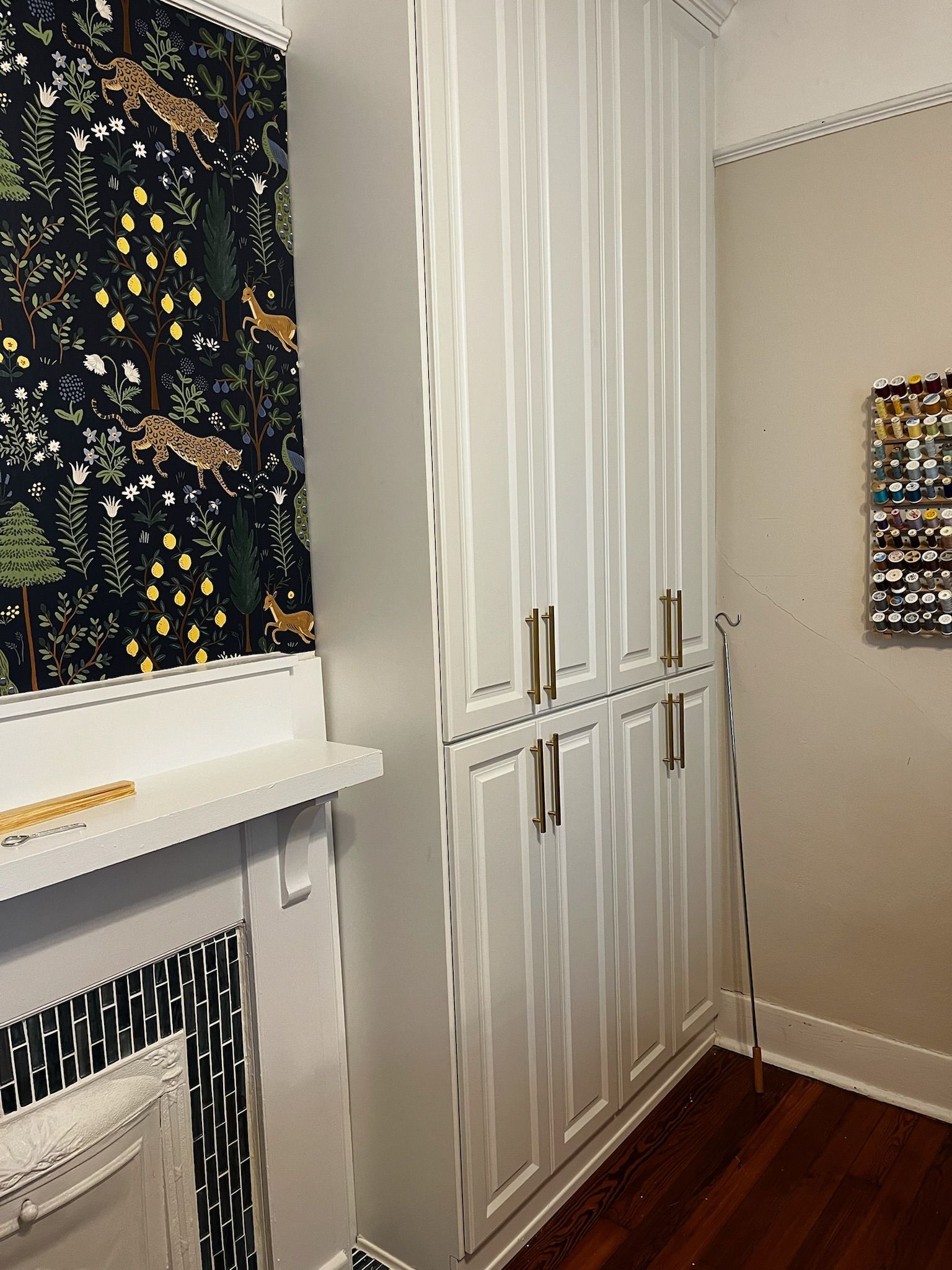 Tall white built-in storage cabinet with gold handles next to a patterned wallpaper and fireplace.
