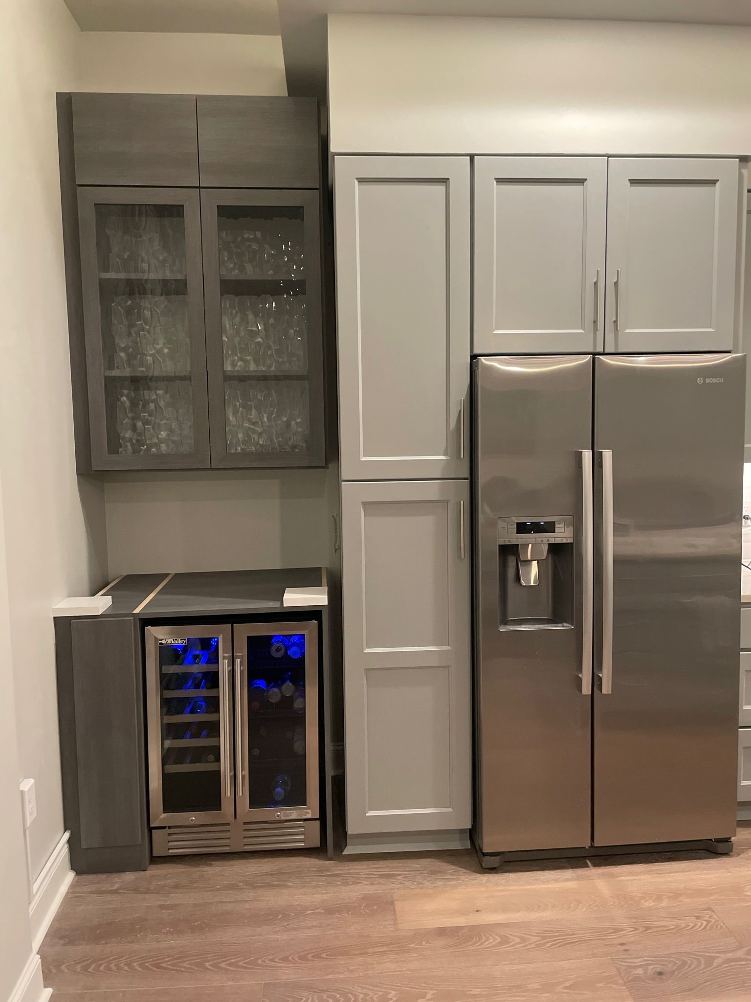 A kitchen area with cabinets and a refrigerator. Includes a wine cooler, gray and stainless steel appliances, and brown flooring.