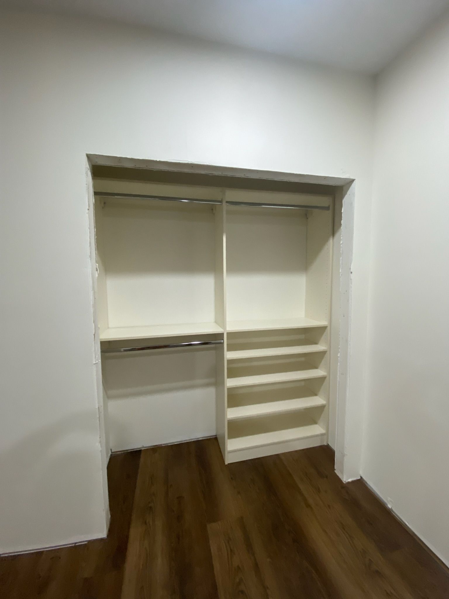 Empty closet with shelves, rods, and white trim against a white wall. Dark wood floor.