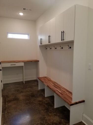 White mudroom with a wood bench, cabinets, and desk against a stained concrete floor.