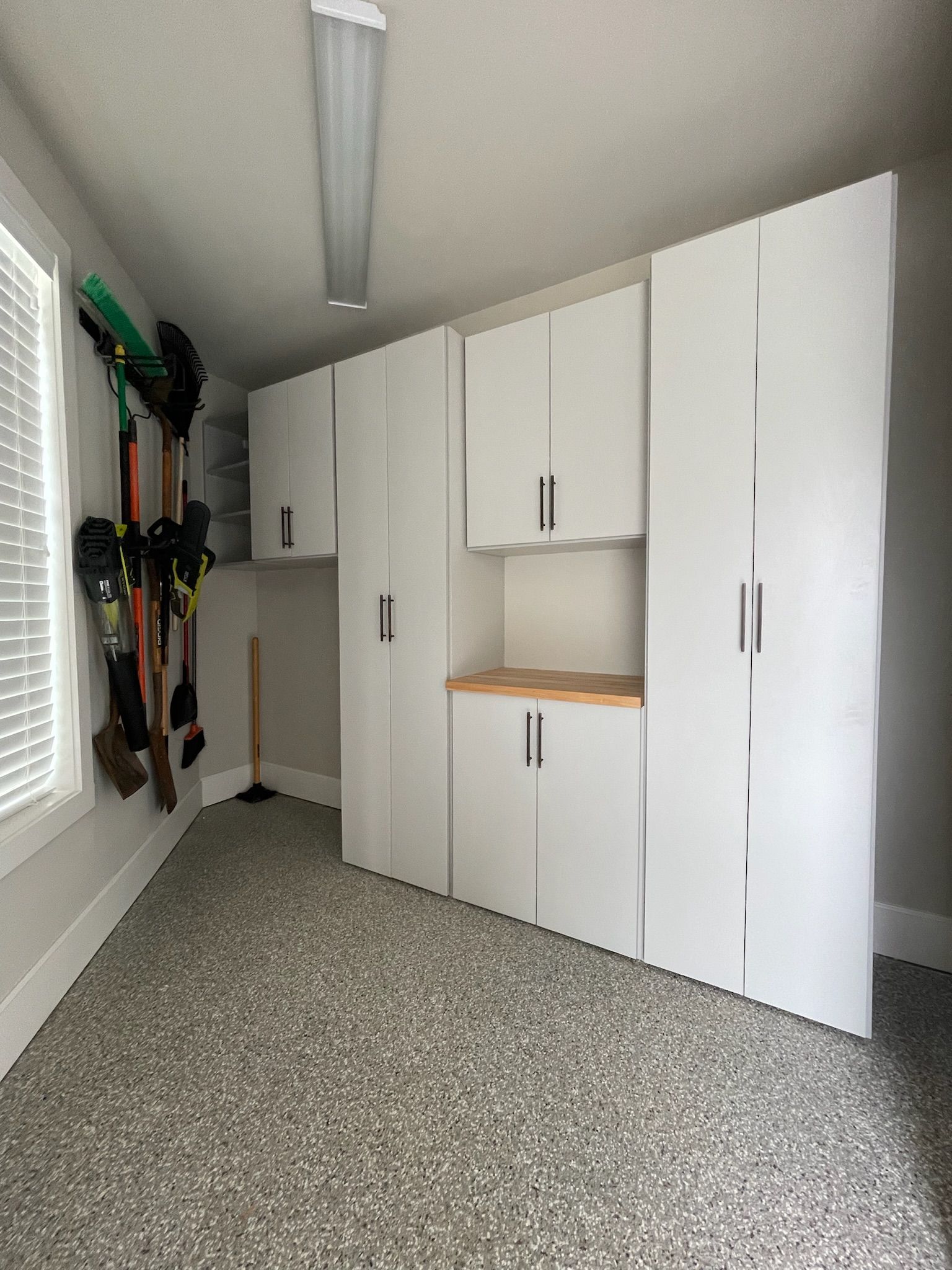 A storage room with a speckled floor, white modular cabinets, and gardening tools hanging on the left wall.