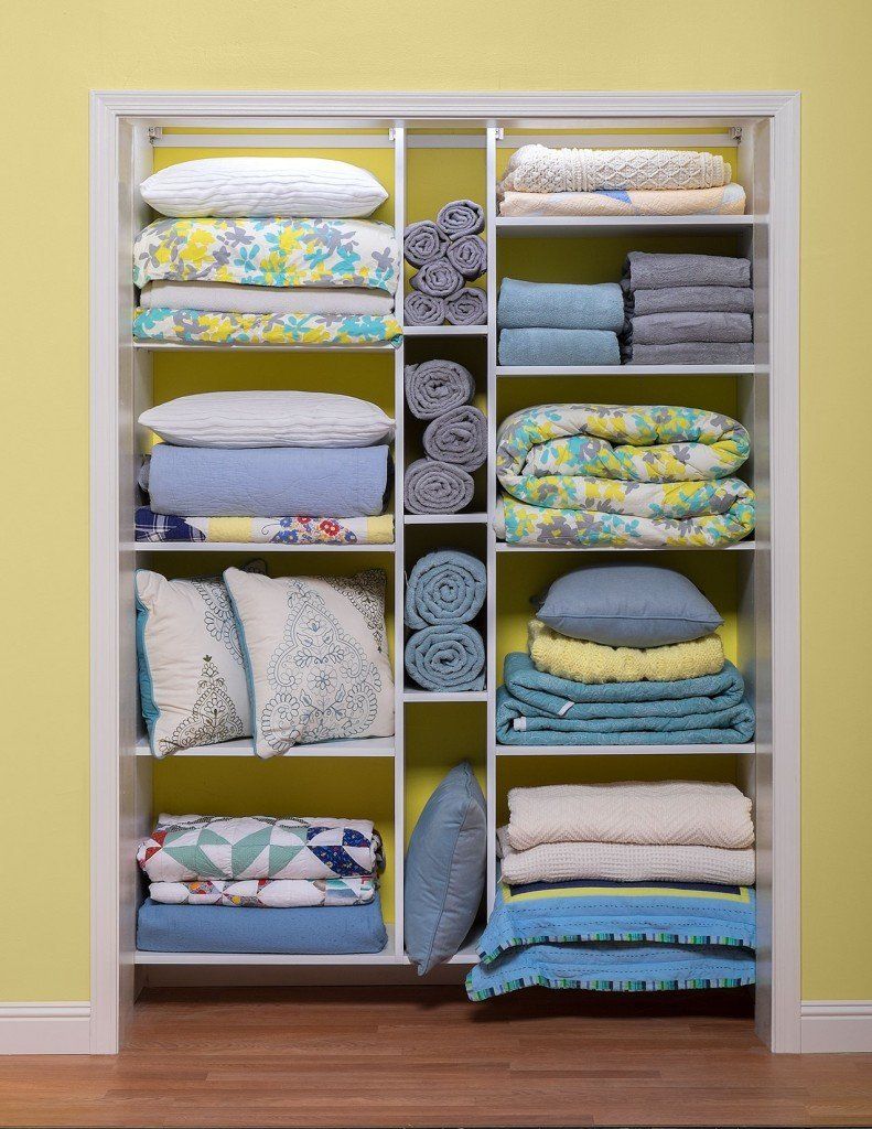 White shelving unit inside a closet, filled with folded blankets, pillows, and towels in shades of blue, yellow, and white.
