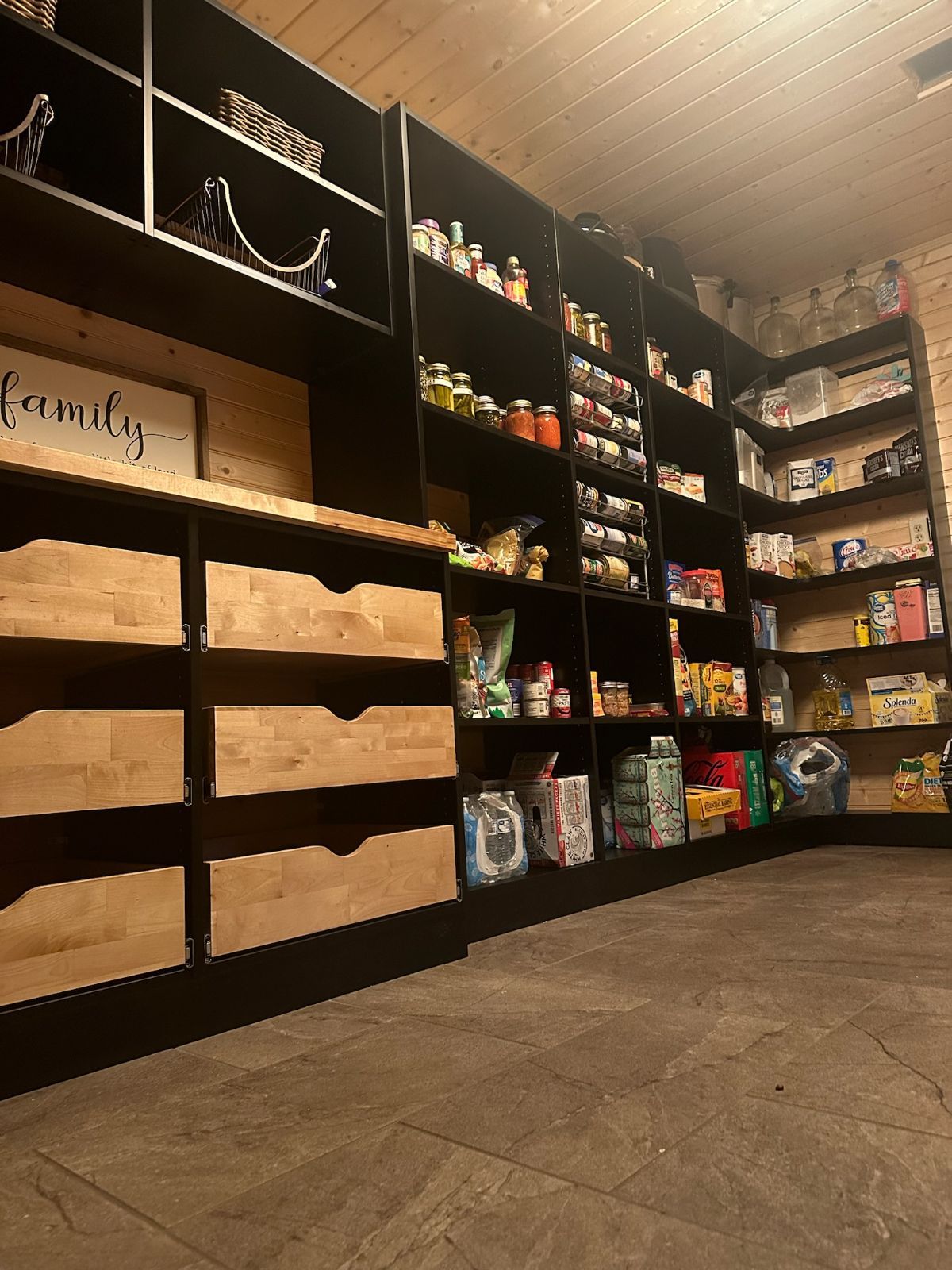 Dark pantry with built-in shelves stocked with food items and wooden drawers. Beige walls and floor.