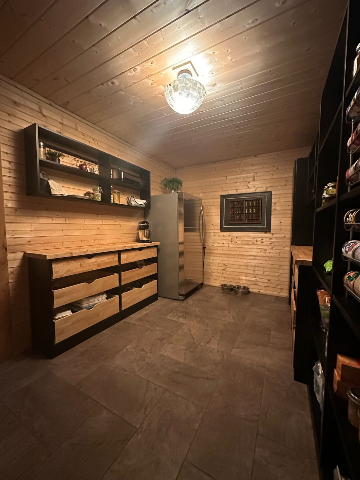 Walk-in closet with wood-paneled walls, dark shelving, drawers, and tile floor, lit by a central light fixture.