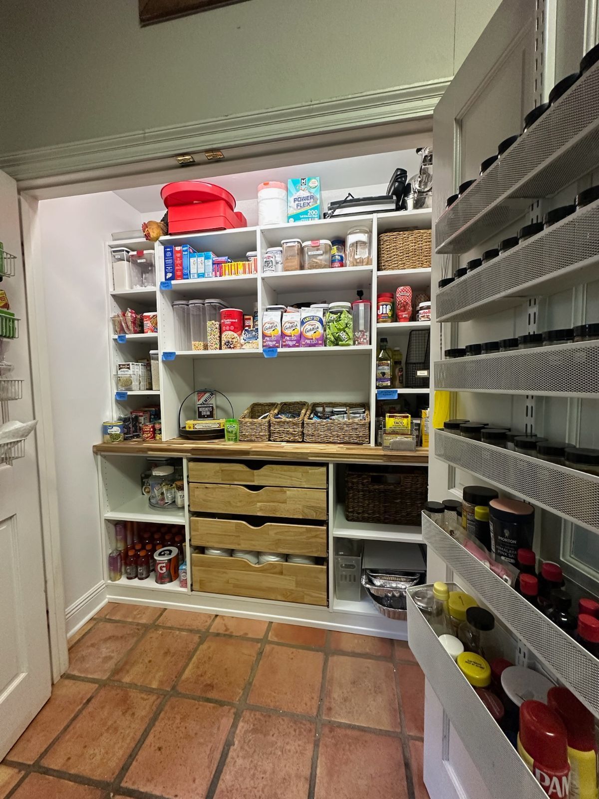 A pantry with white shelves filled with food items, a wooden countertop, and spice racks on the right.