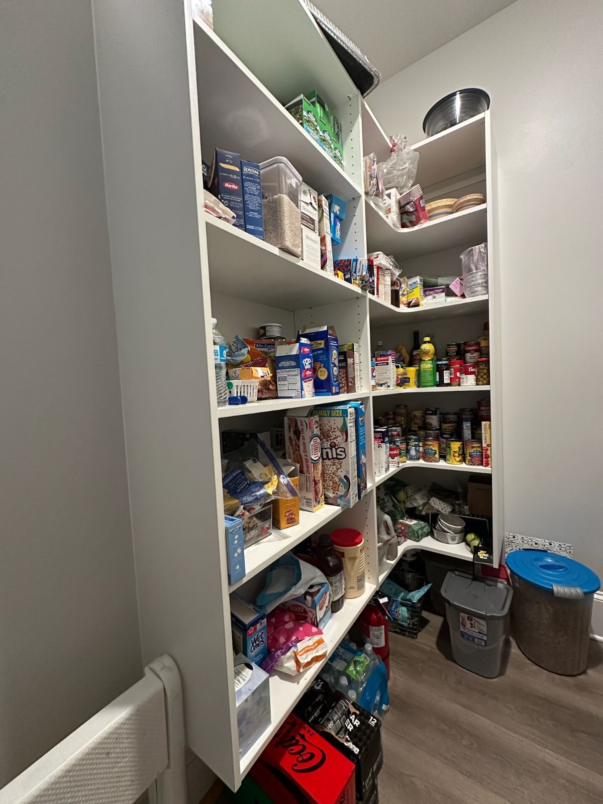 White pantry shelving unit filled with food items in a corner, with storage bins on the floor.