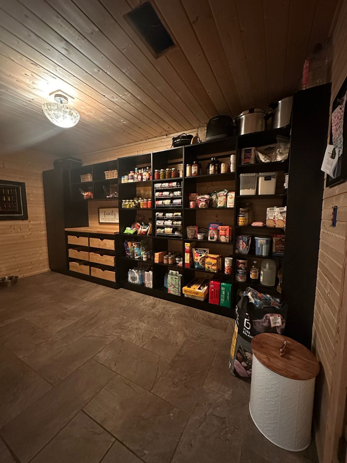 Pantry with black shelving, assorted food items, wooden ceiling, and a dog bowl on the floor.