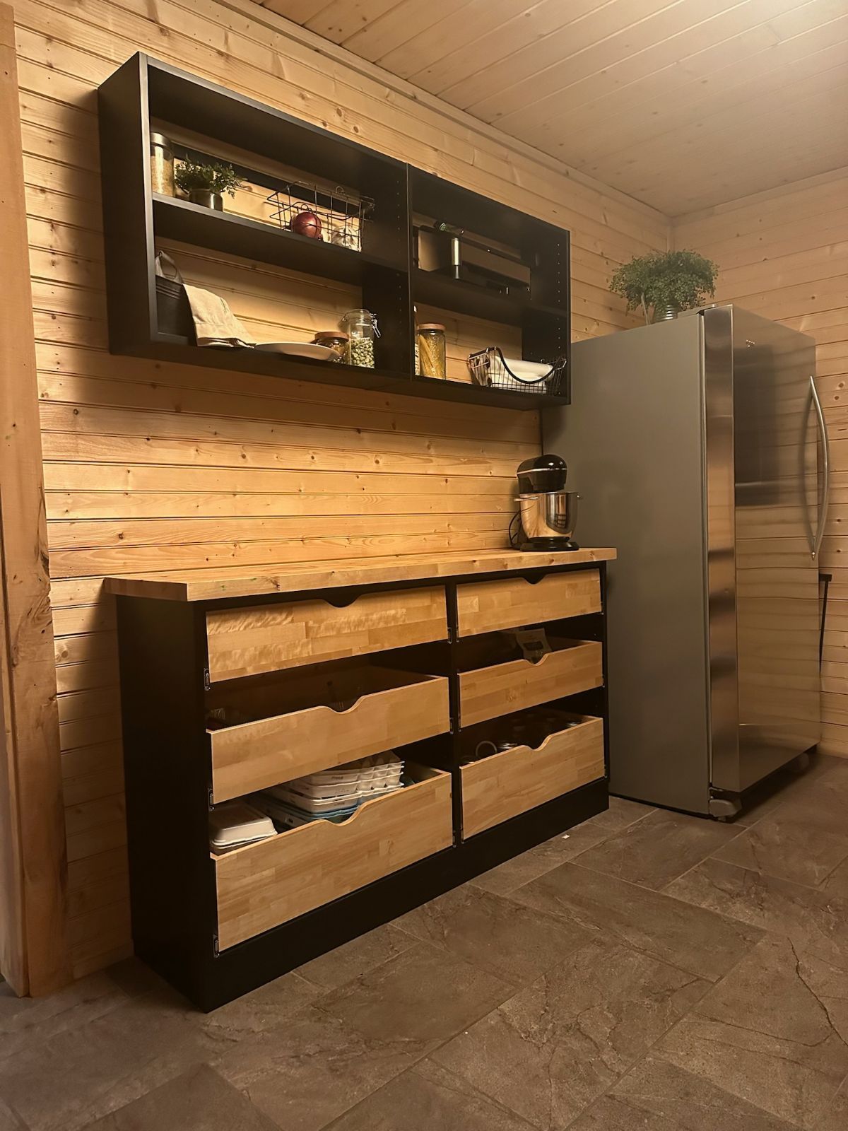 Wooden cabinet with drawers and black shelves on wood-paneled wall, next to a stainless steel refrigerator.