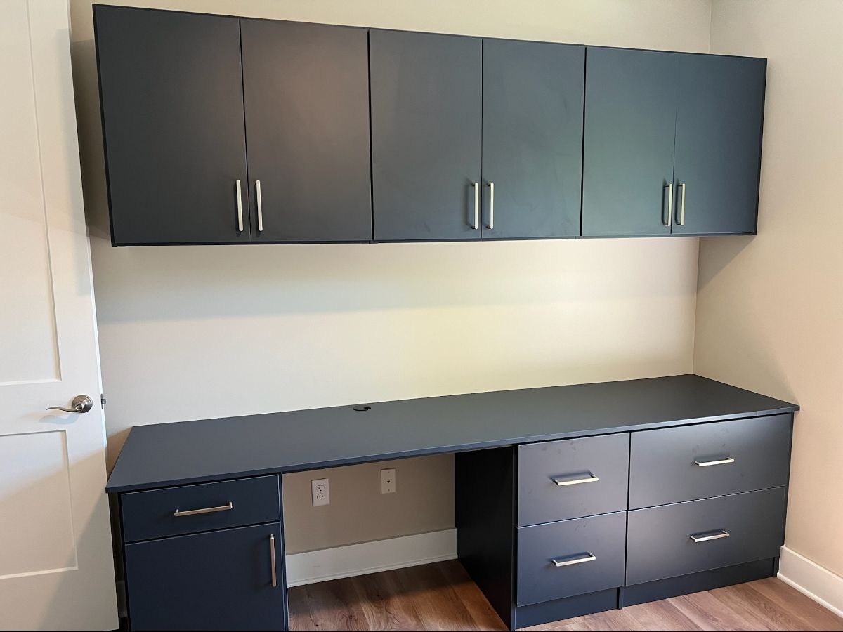 Built-in dark gray desk and cabinets. Overhead cabinets above desk with silver handles. Beige wall, wooden floor.