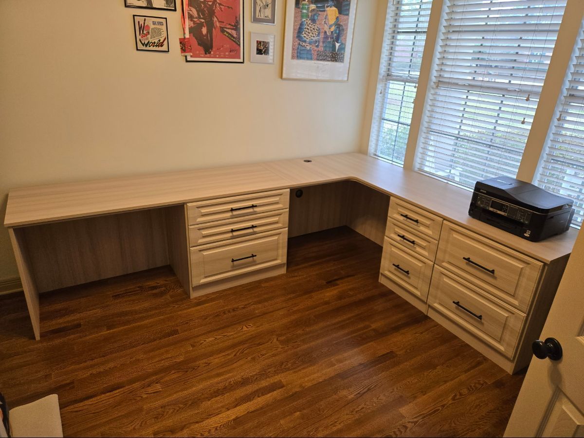 L-shaped desk with drawers by a window and a printer. Wooden floor and posters on wall.