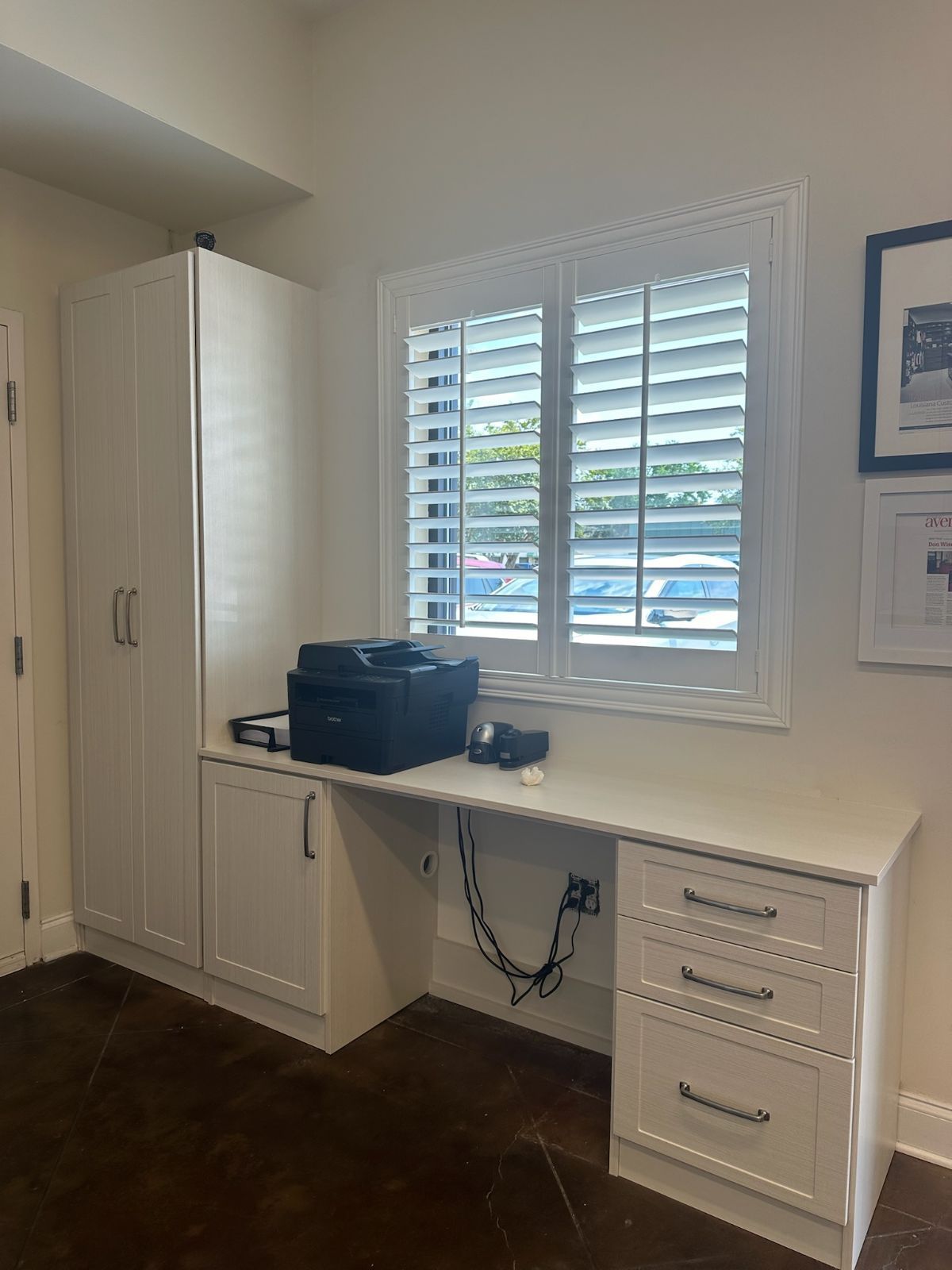 Built-in desk with printer and storage cabinets next to window with shutters. Dark wood floor.
