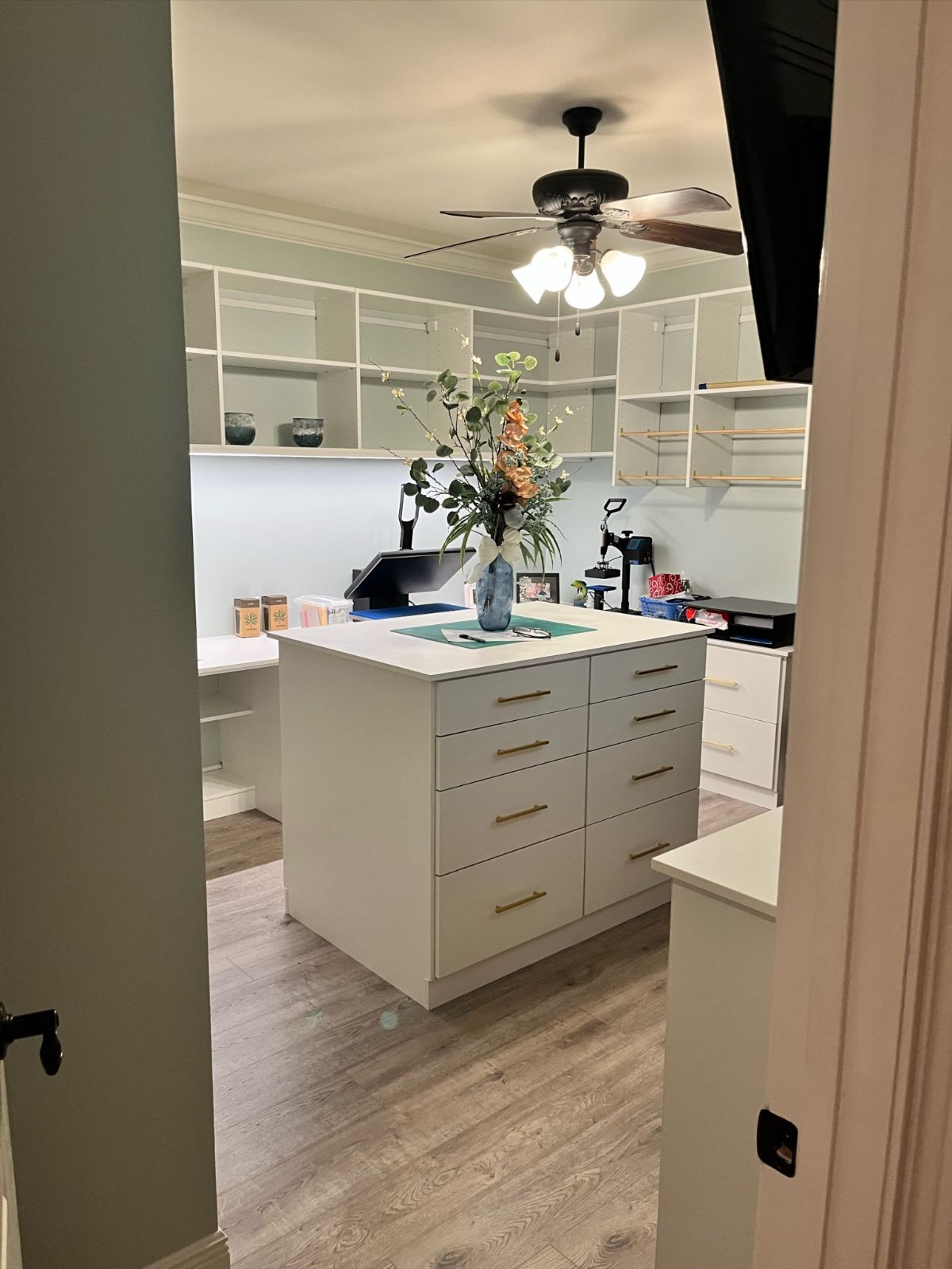 A craft room with white cabinets, a central island, and shelving. A vase of flowers sits on the island.