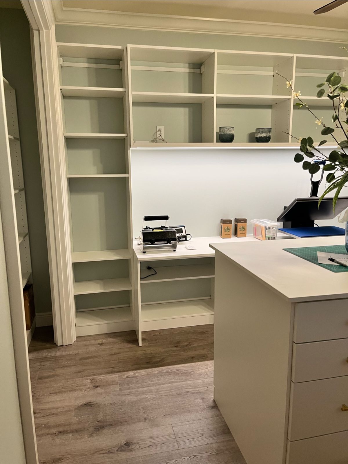 White built-in shelving and desk, pale green walls, wooden floor, and a white cabinet.