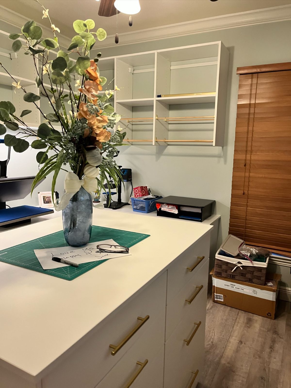 White craft table with flowers, shelves, and printer in a light blue room.
