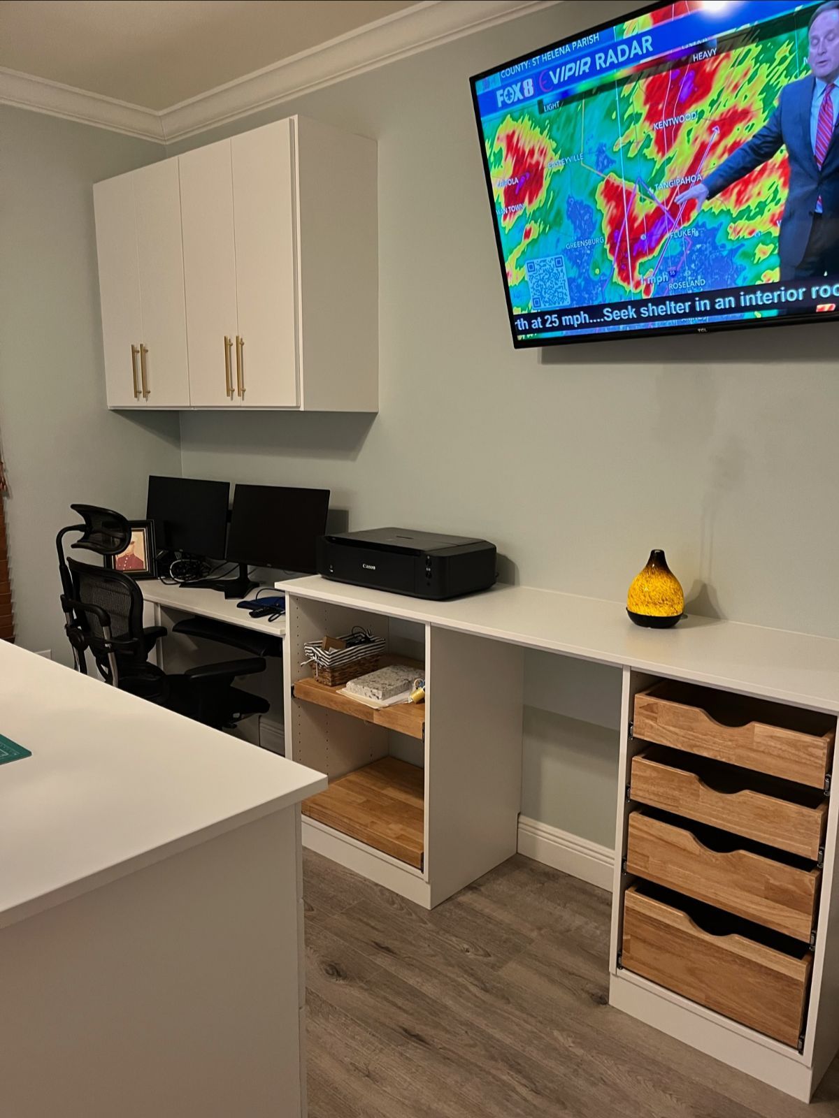 Home office with desk, overhead cabinets, TV, and wooden drawers. Monitors, a printer, and a chair.