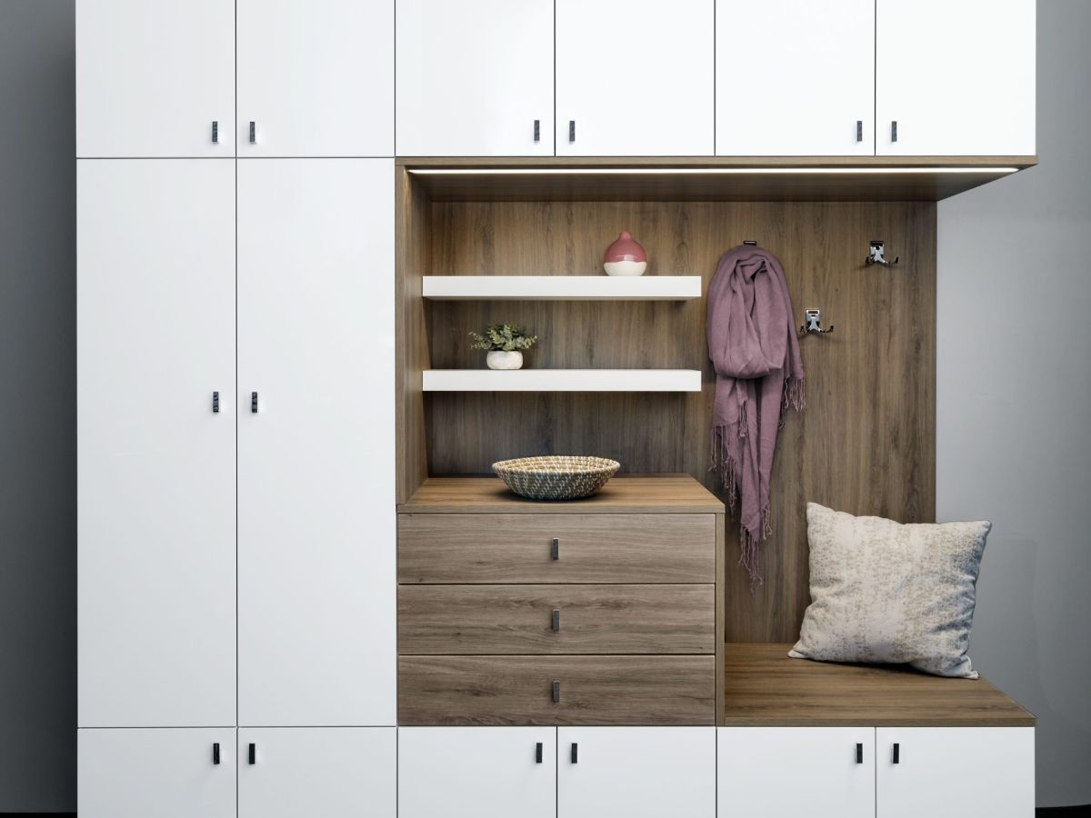 White and wood entryway unit with storage cabinets, bench, and coat hooks. A pillow and scarf rest on the bench.
