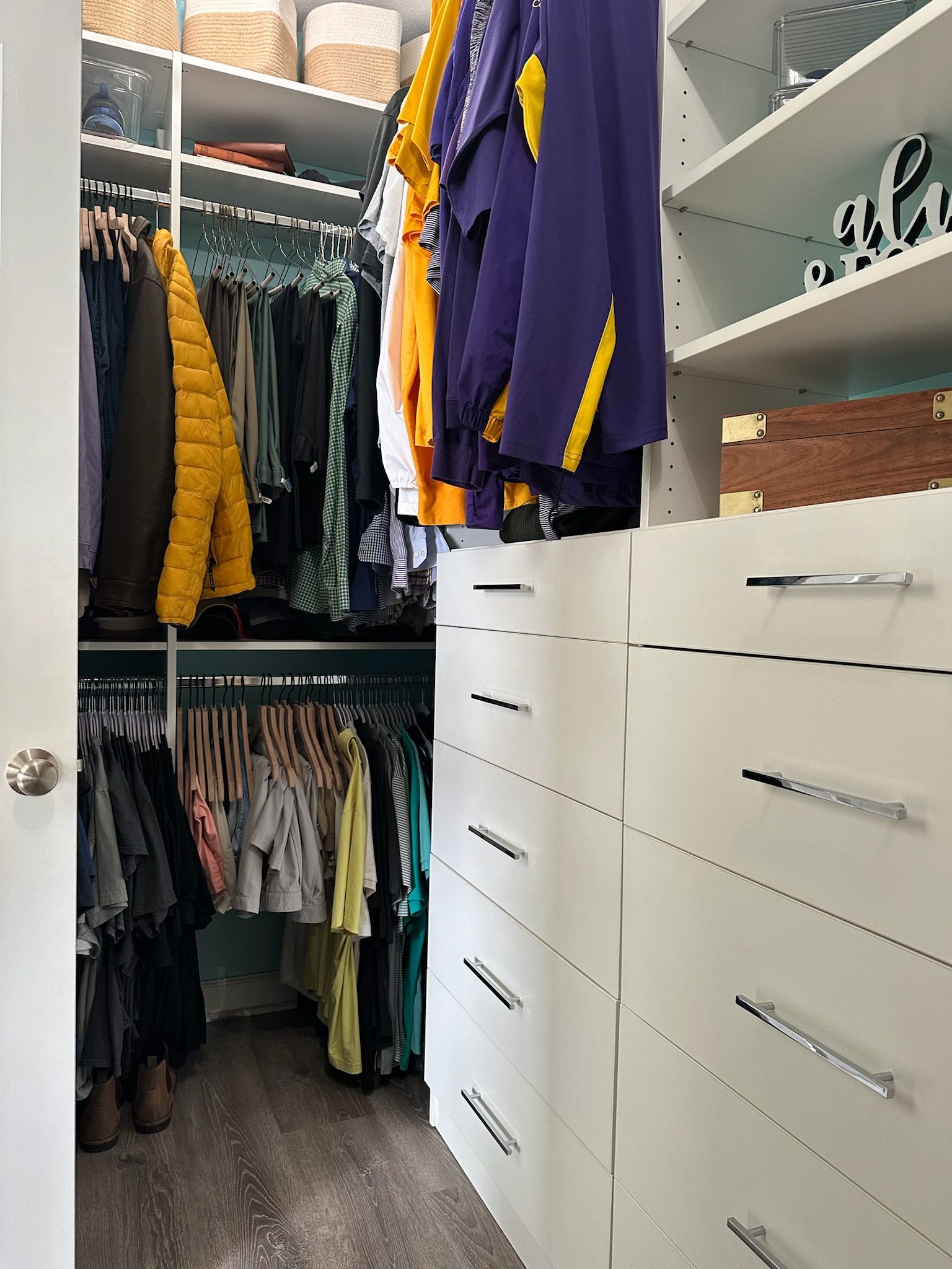 Walk-in closet with white shelves, drawers, and hanging clothes.  Hanging clothes are different colors.