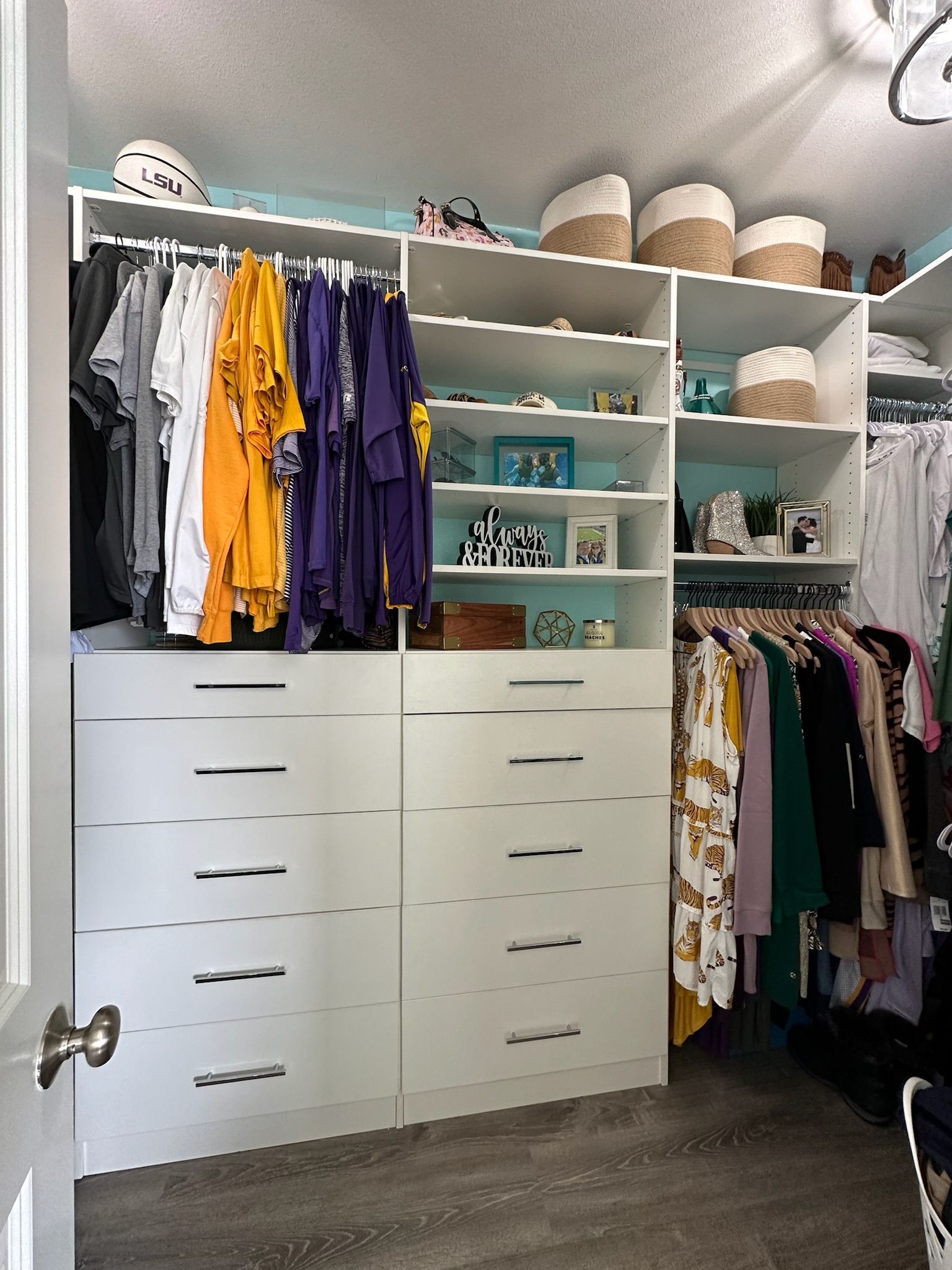 A well-organized white closet with hanging clothes, drawers, shelves with baskets and decor, and a door.