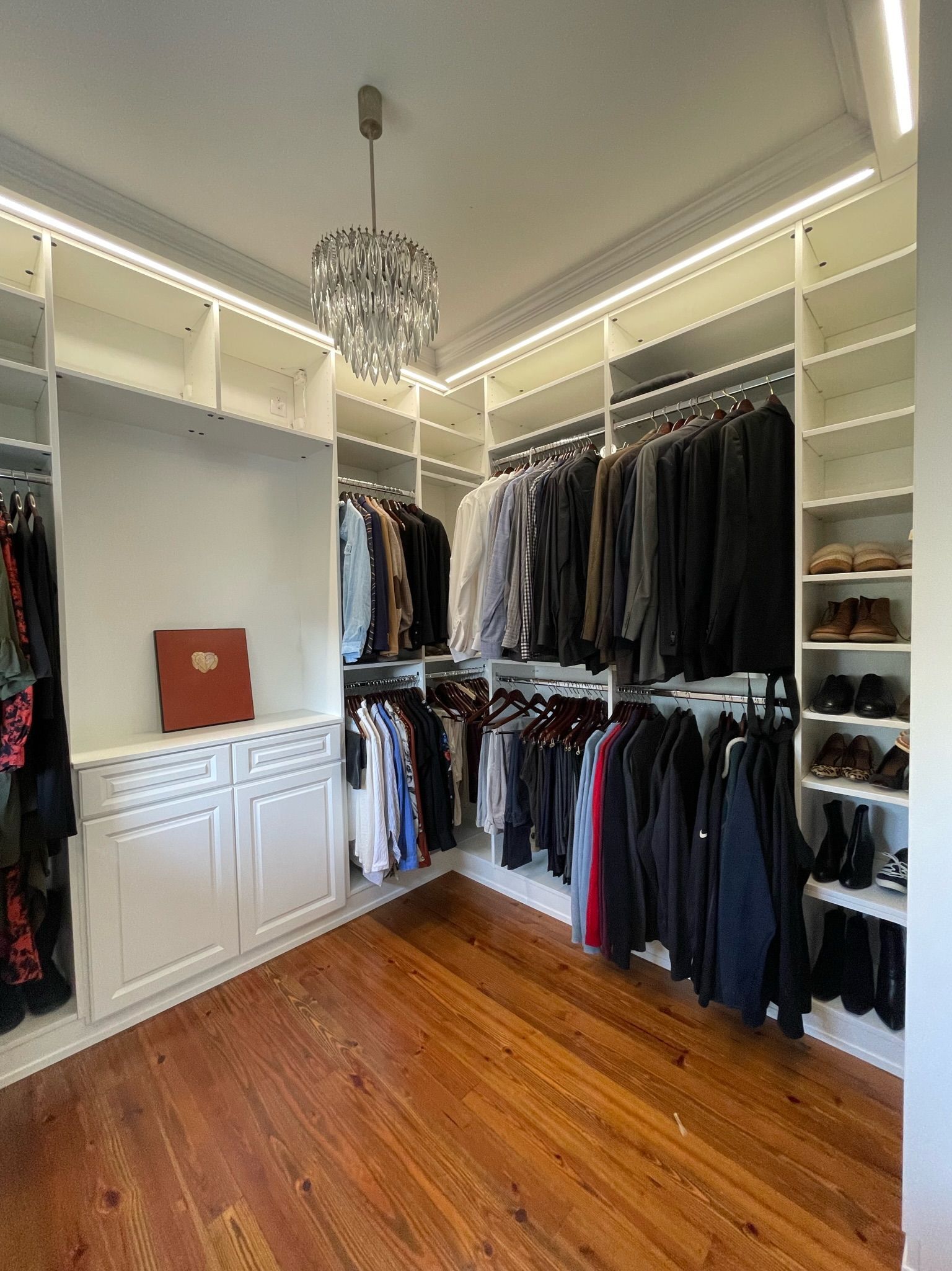 Walk-in closet with white shelves and wood floor, containing hanging clothes and shoes. A chandelier hangs from the ceiling.