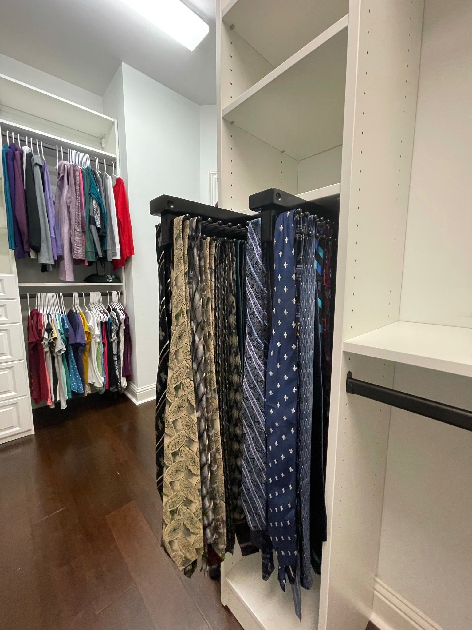 Walk-in closet with hanging clothes. Tie rack with patterned ties, dark wood floor, and white shelves.
