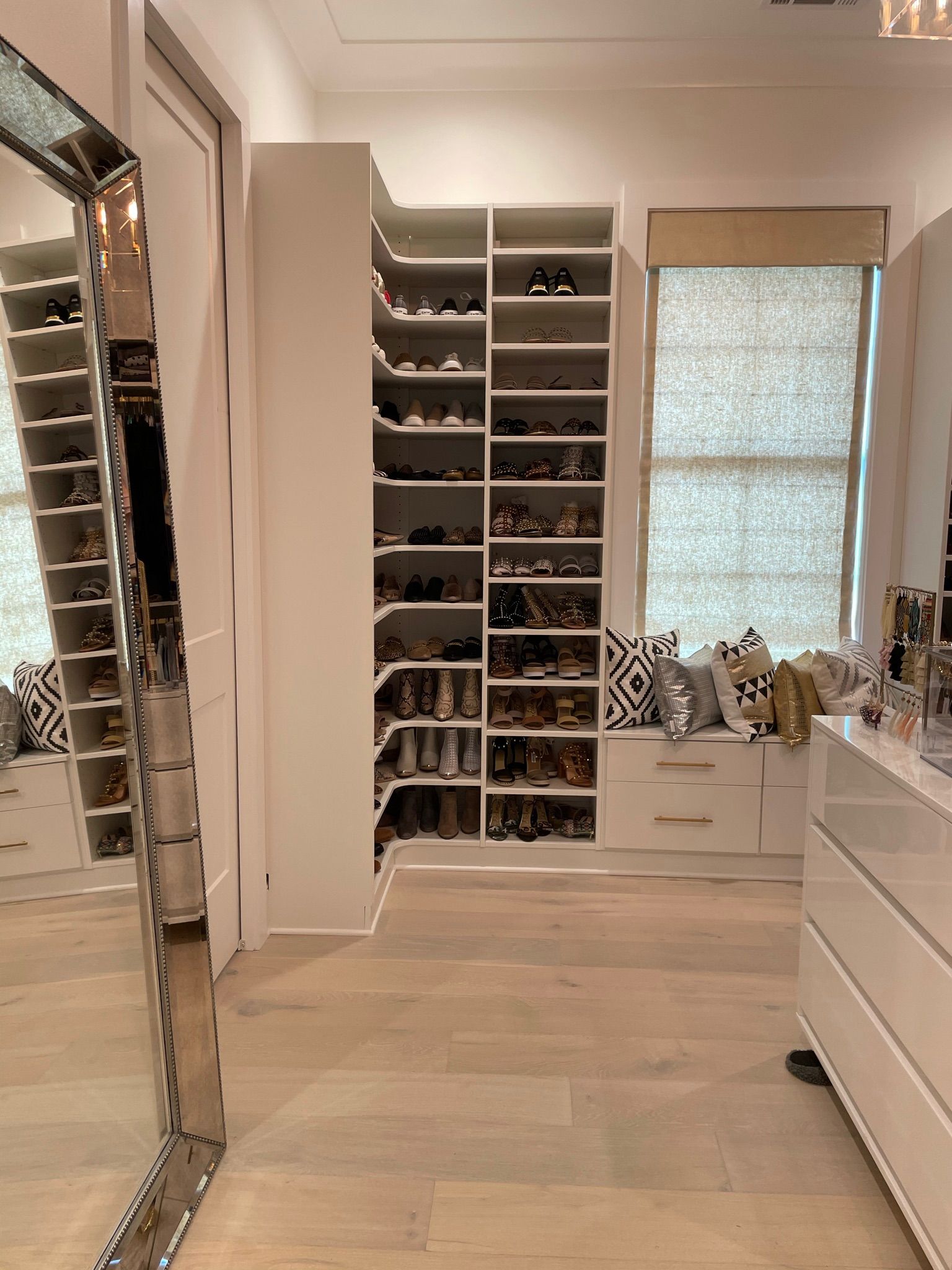 Walk-in closet with shoe shelves, a large mirror, window seat, and dresser; light wood floors, neutral colors.