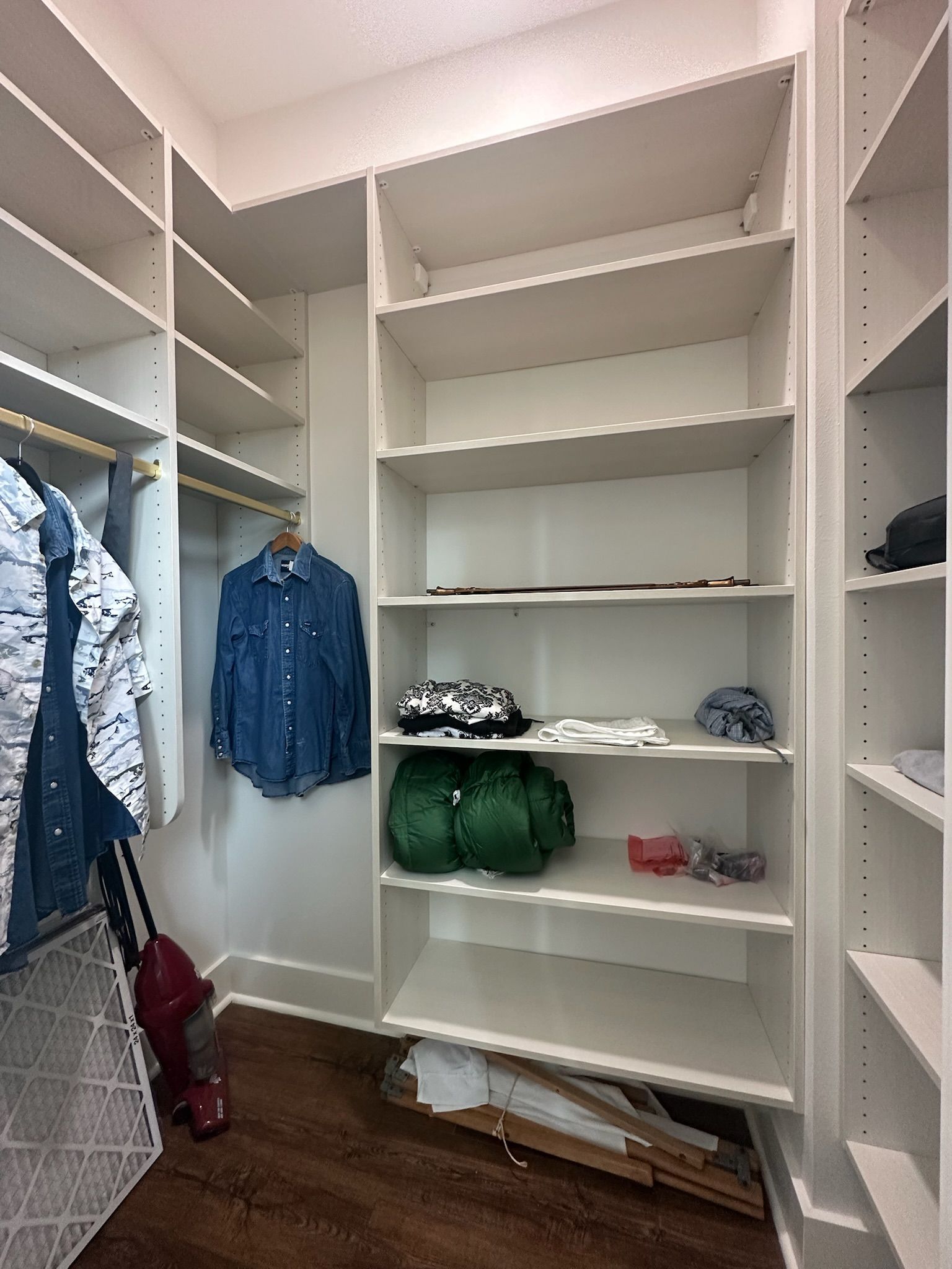 Empty white closet with shelves and hanging rods, some clothes, and a vacuum cleaner.