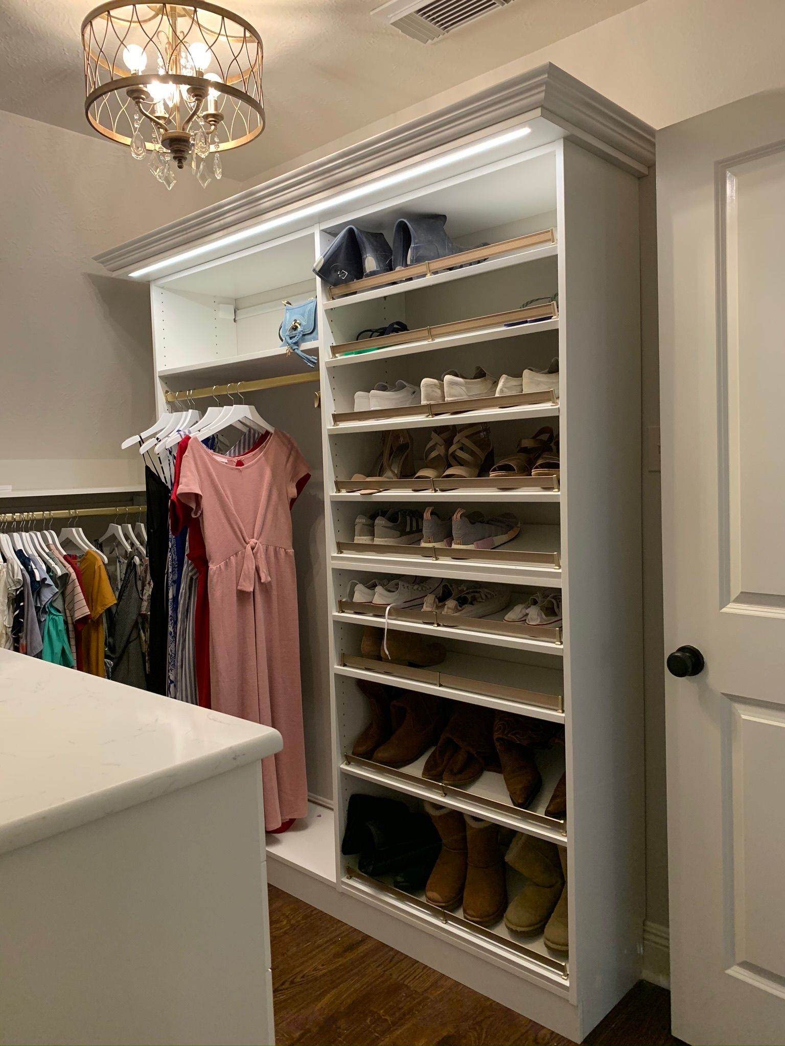 Walk-in closet with built-in shoe shelves and hanging clothes. White cabinets, wooden floors, and chandelier lighting.