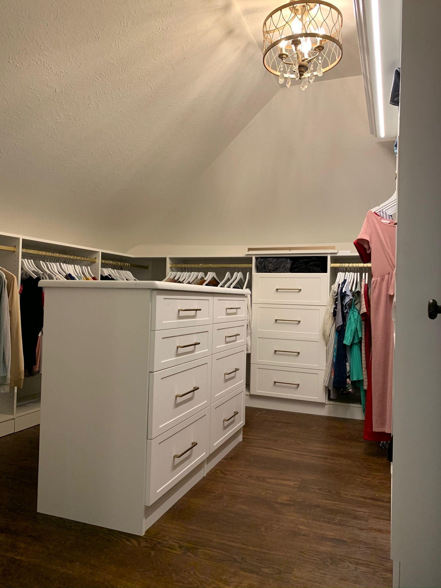 Walk-in closet with white drawers and hanging clothes. Warm-toned wood floor and ceiling light.