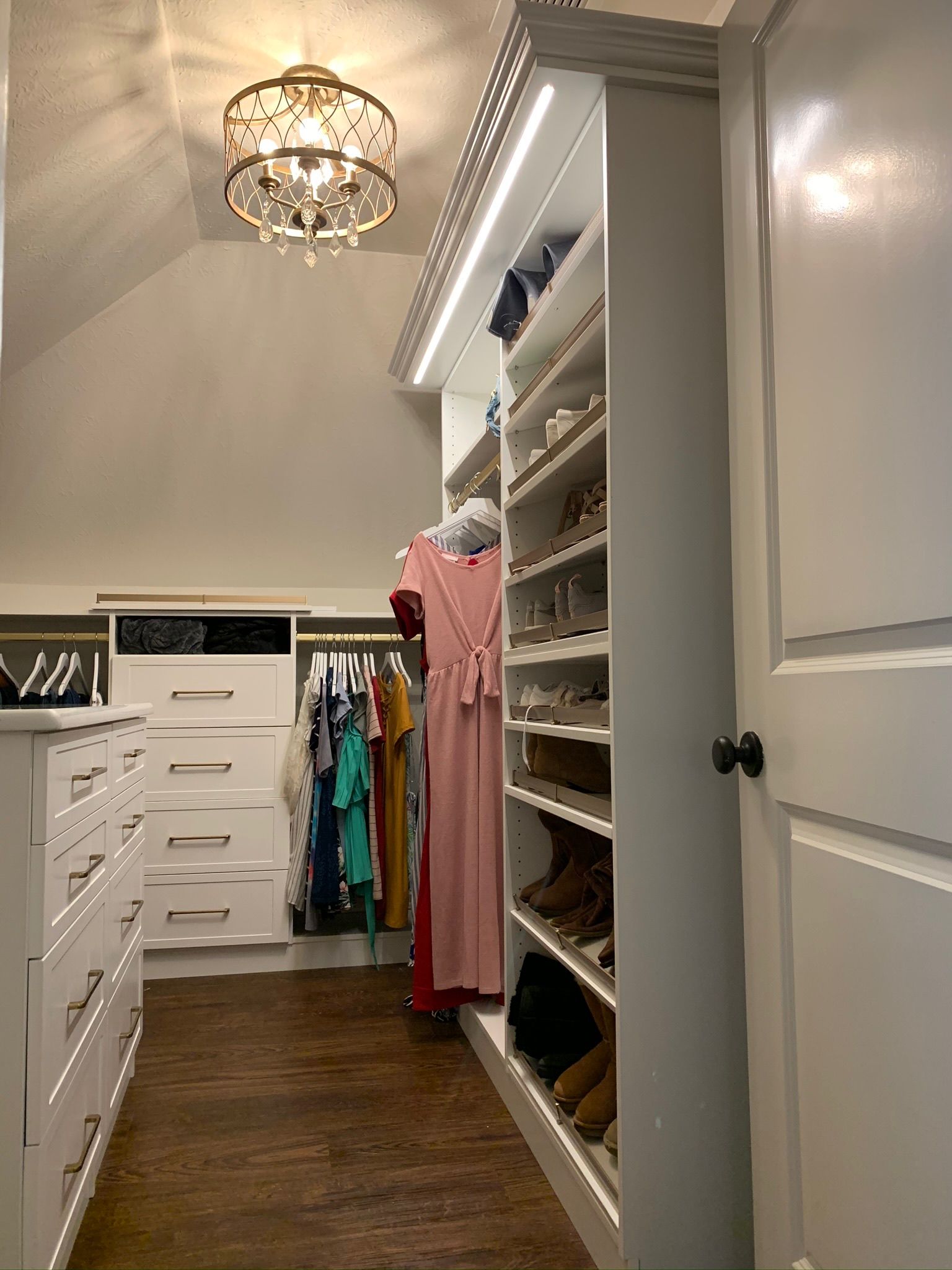 A walk-in closet with white cabinetry, built-in shoe shelves, hanging clothes, and a chandelier; brown floors.