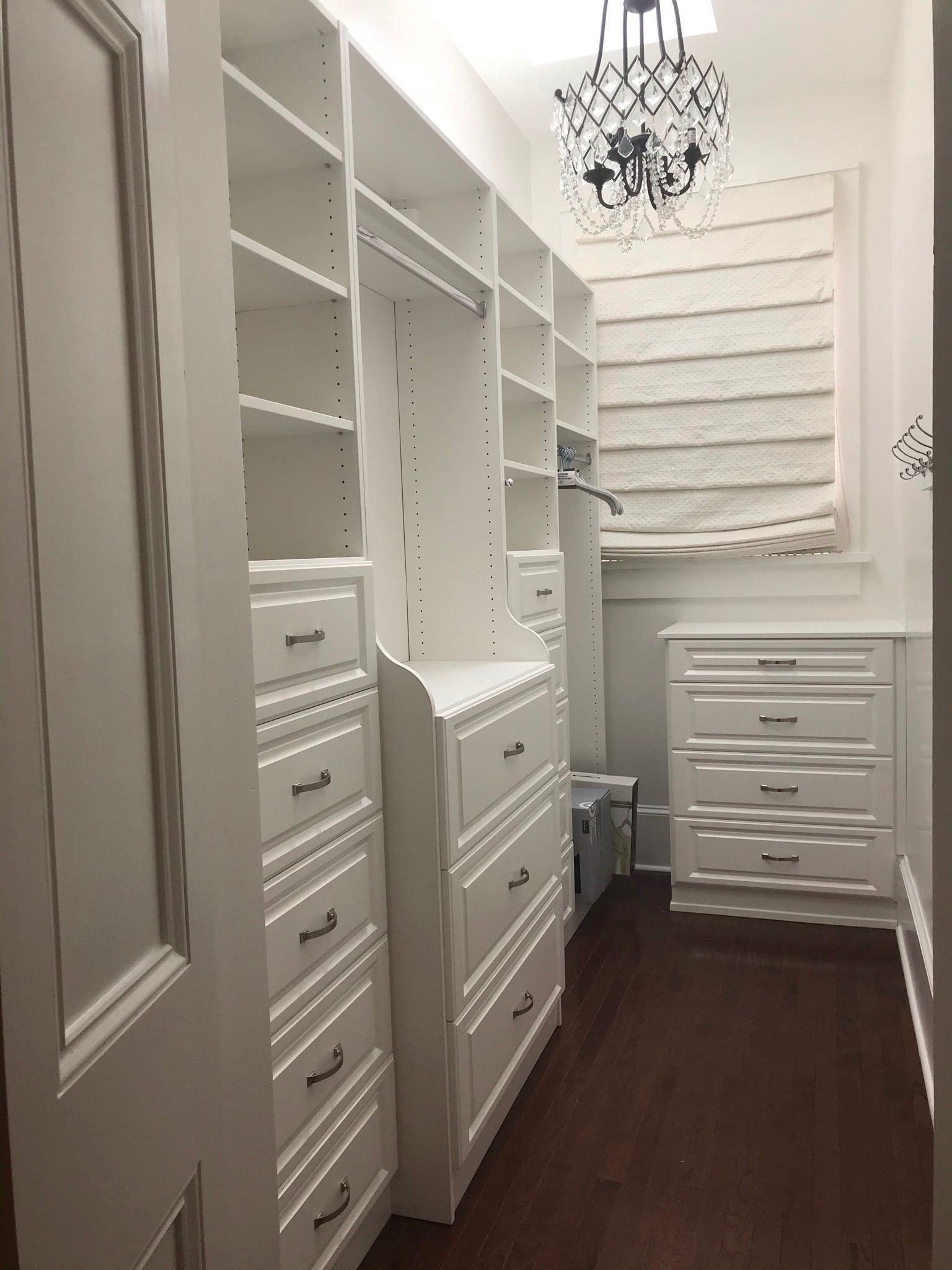 White custom closet with drawers and shelves; dark wood floor, chandelier, and a window with a roman shade.
