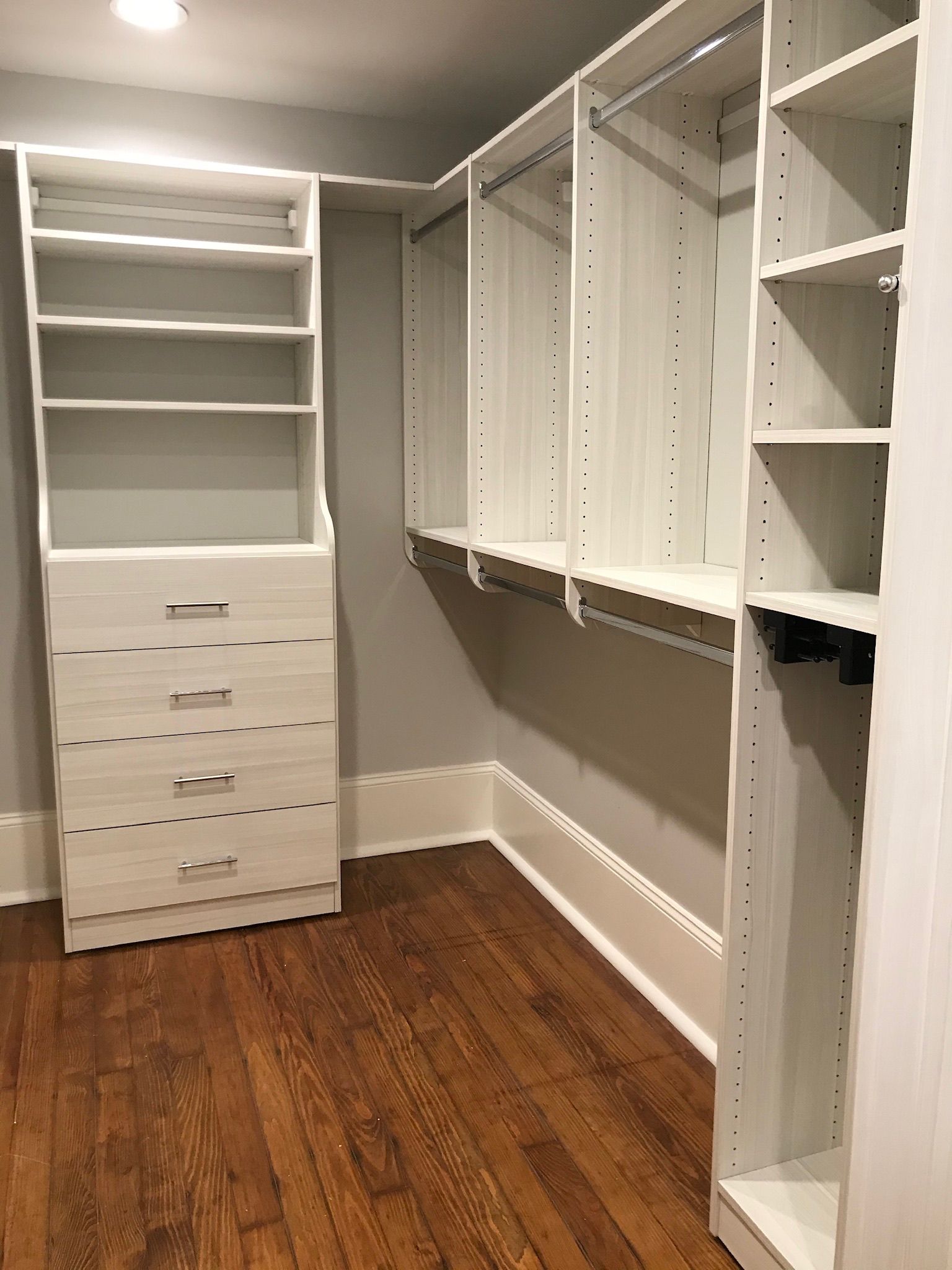 A walk-in closet with white shelving, drawers, and wooden floor. Gray walls.