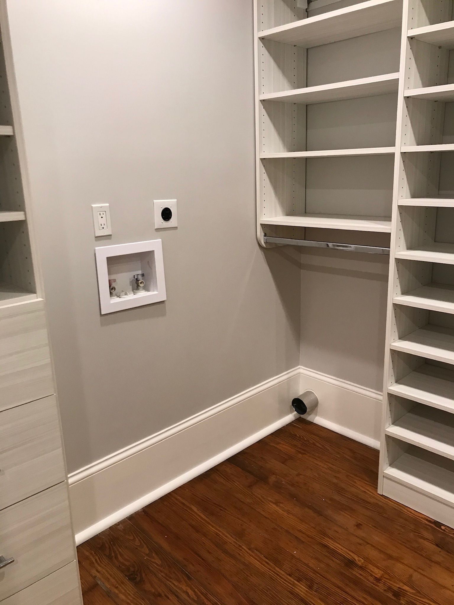 Laundry room with shelves, electrical outlets, and water hookups on gray wall; wood floor.