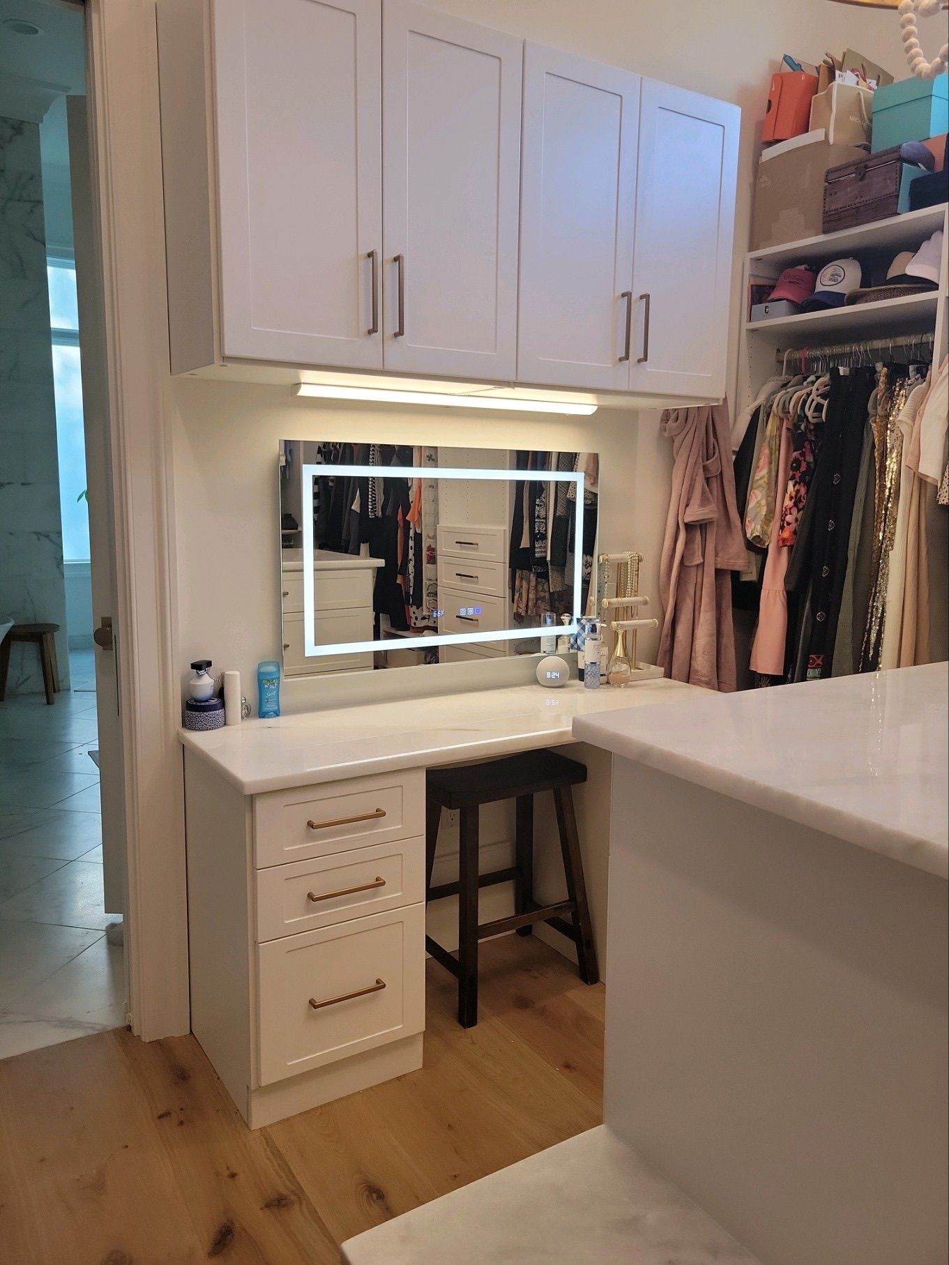 Vanity area in a walk-in closet; white cabinetry, marble countertop, lighted mirror, and black stool.