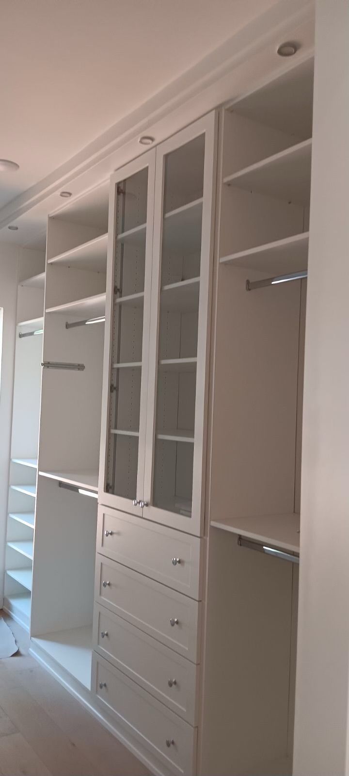White built-in closet with shelves, drawers, and glass-paneled doors. Recessed lighting in the ceiling.