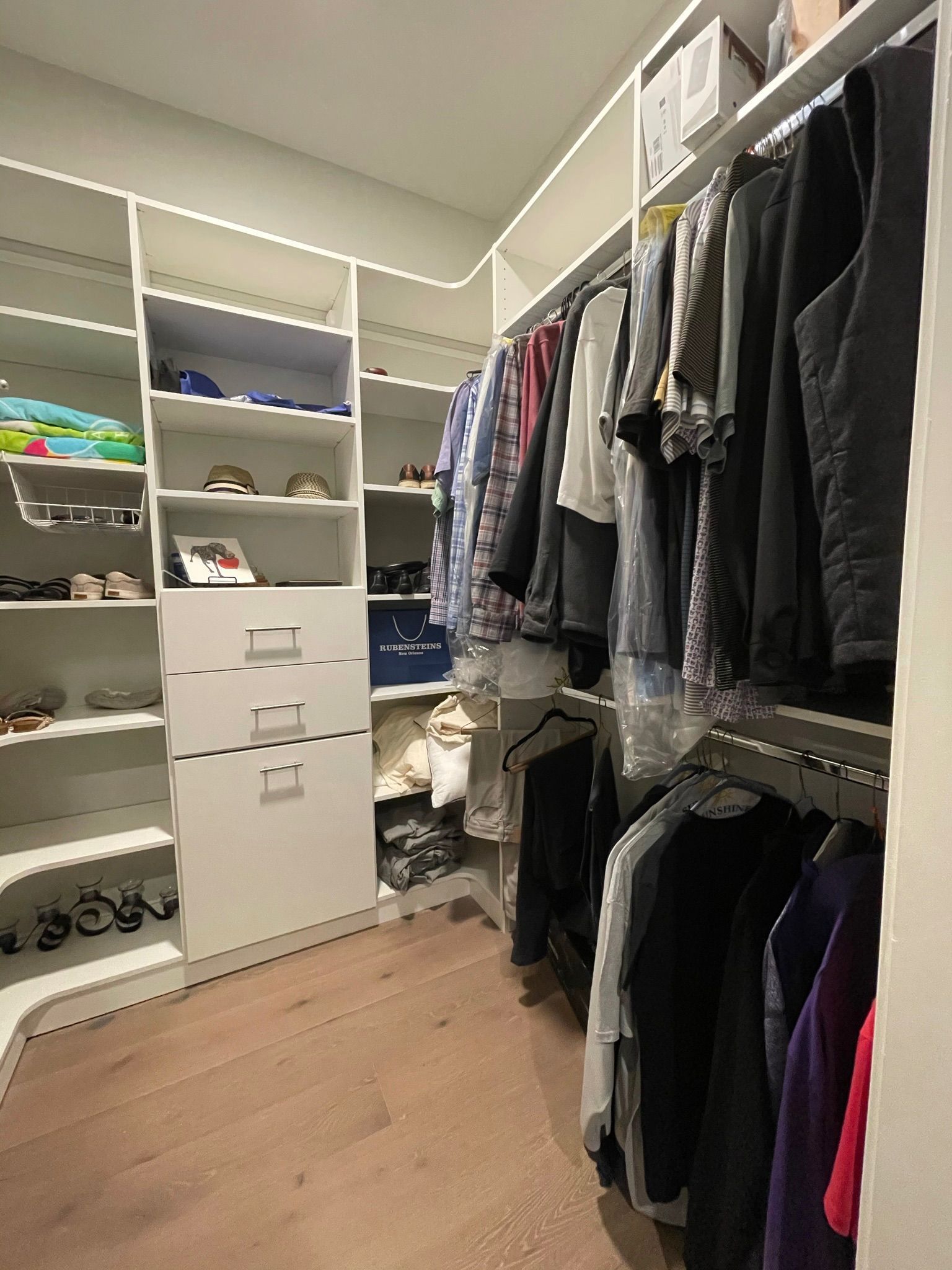 Walk-in closet with white shelving and drawers, clothes hanging on rods, and wooden floor.