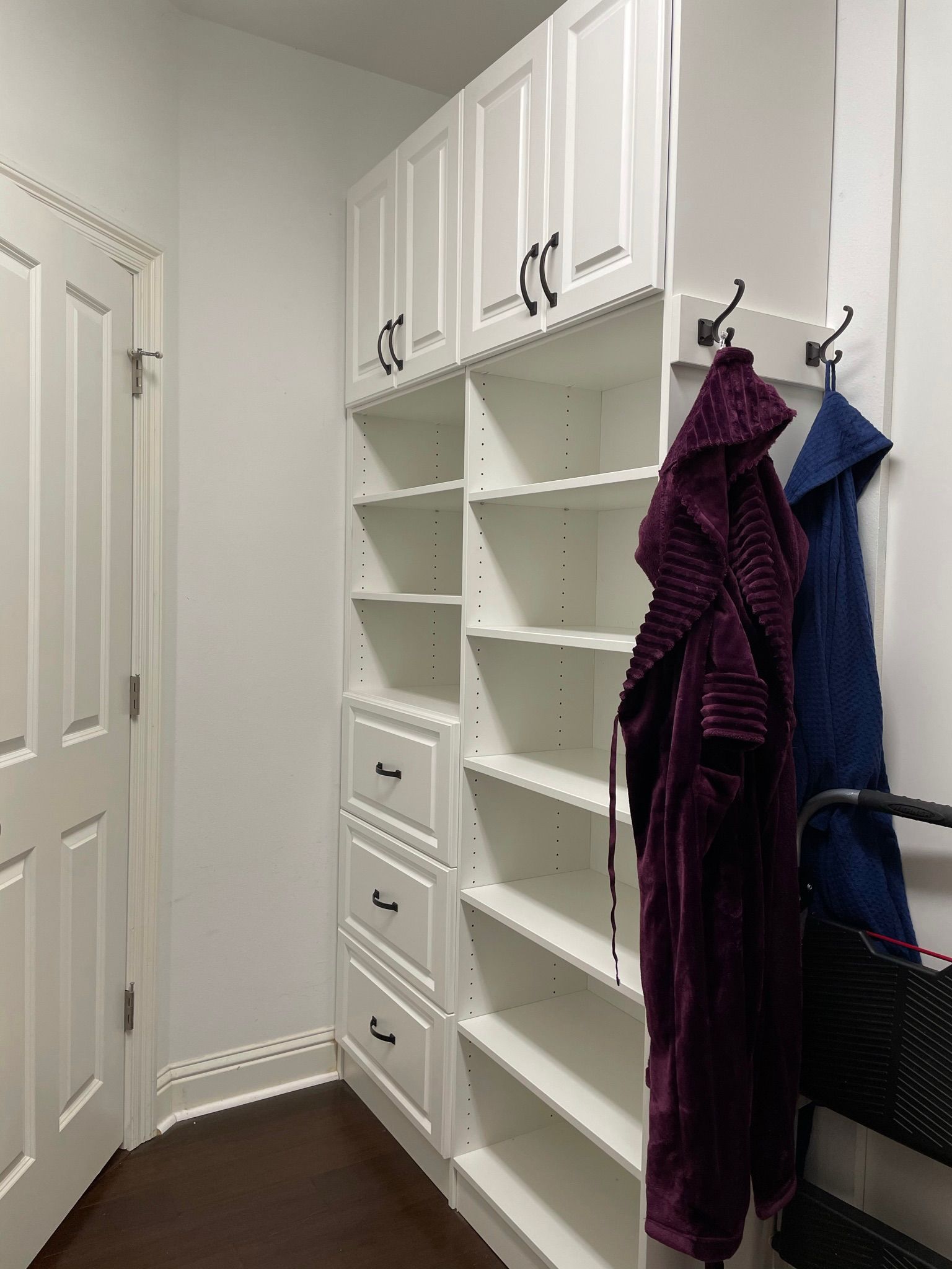 White built-in storage unit with shelves, drawers, and upper cabinets in a hallway. Two robes hang on hooks.