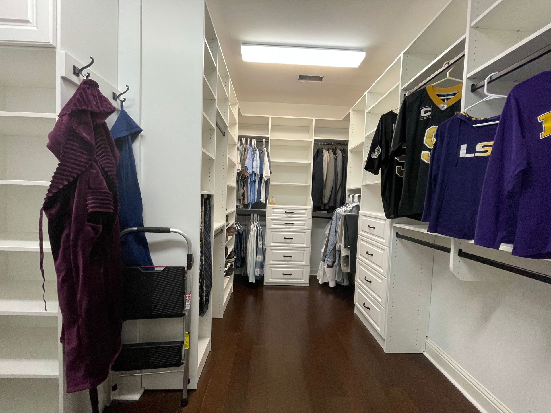 Walk-in closet with white shelving and dark wood floor; robes and clothes hang on racks.
