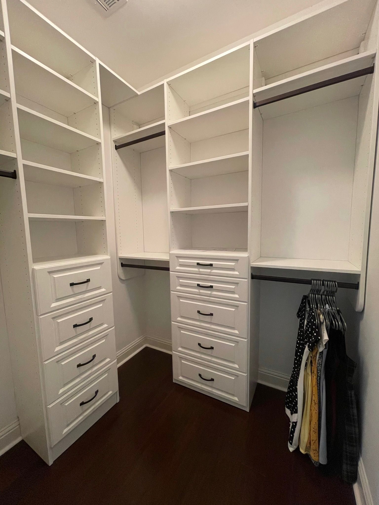 Empty white closet with shelves, drawers, and hanging rods. Dark brown floor.